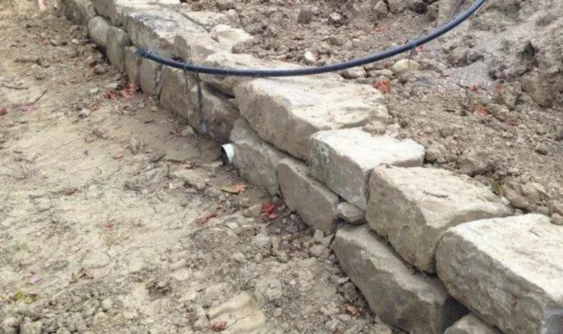 Stone retaining wall under construction along a dirt path, with a blue hose lying across the top.