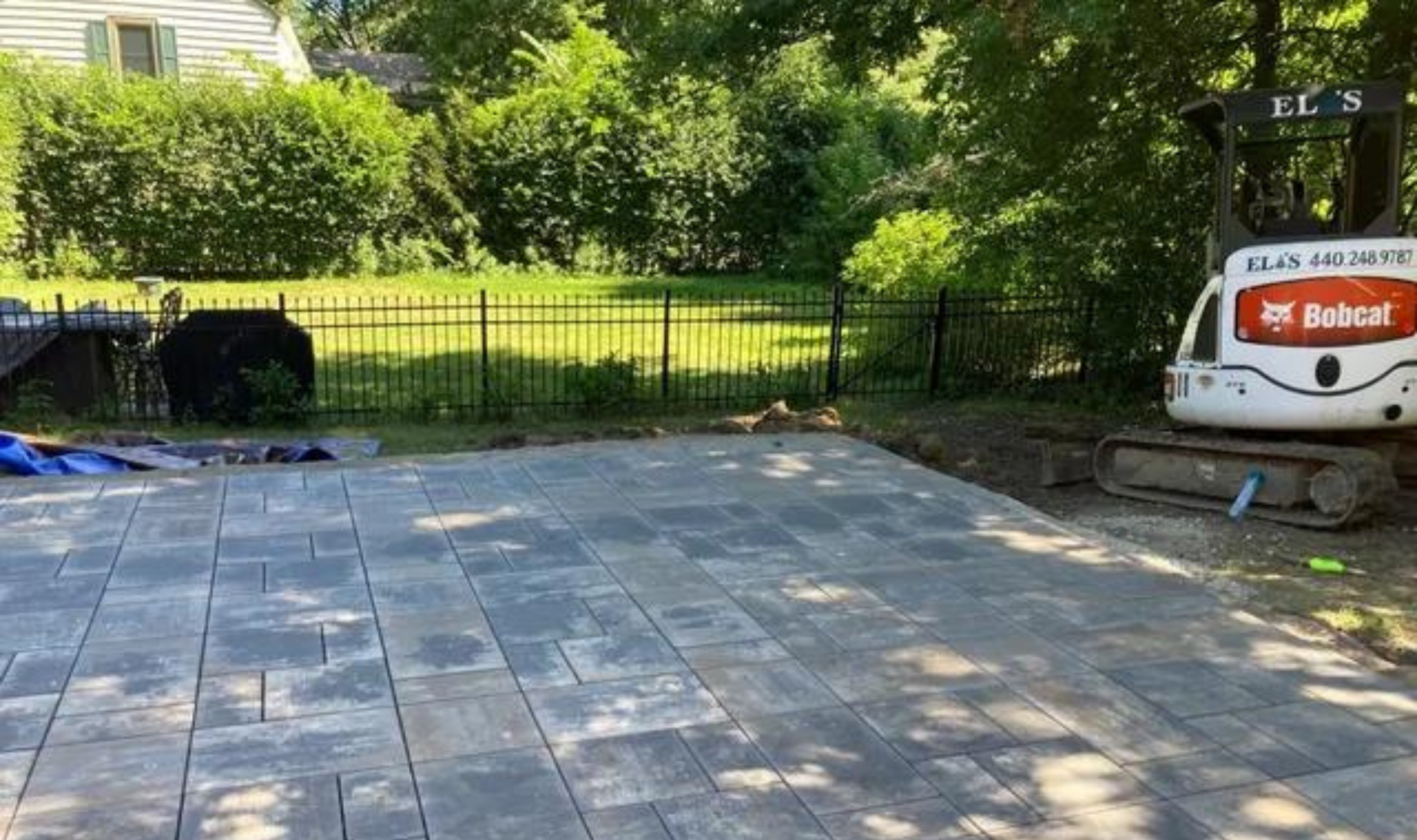 Paved backyard patio with black fence, green lawn, trees, and a Bobcat skid steer at right.