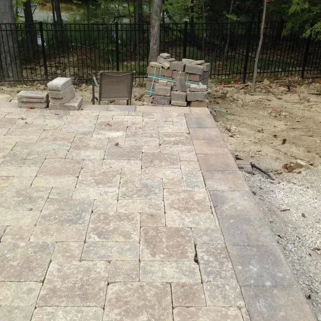 Stone patio under construction beside a fence, with pavers, stacked blocks, and dirt edge