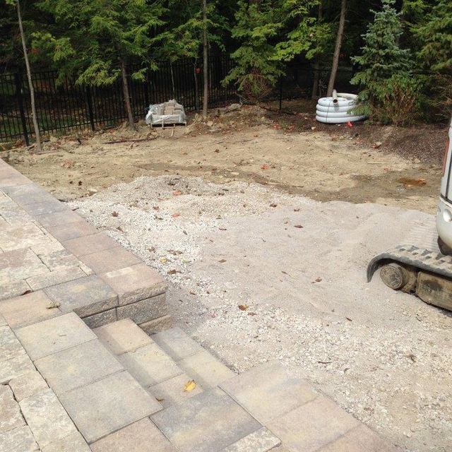 Backyard construction area with concrete pad, paver steps, trees, and utility lids along the fence