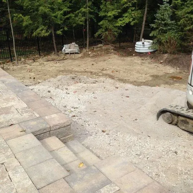 Empty gravel area beside a paver patio, bordered by trees and a metal fence.