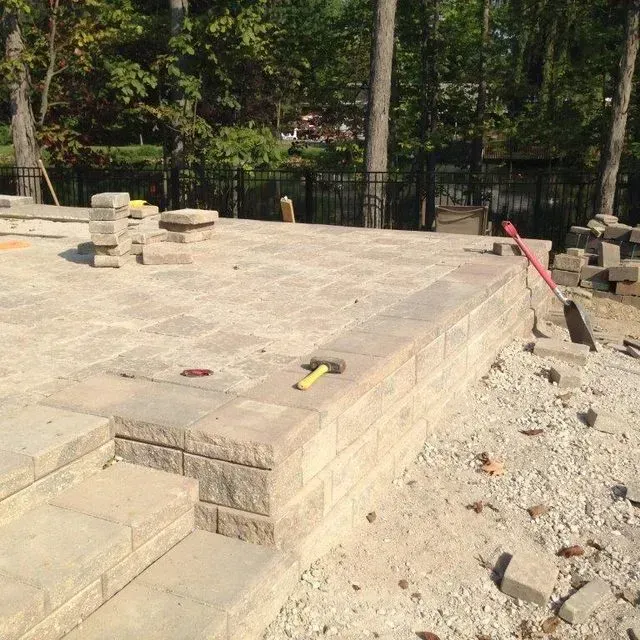 Stone patio under construction with steps, tools, and scattered blocks beside a wooded area