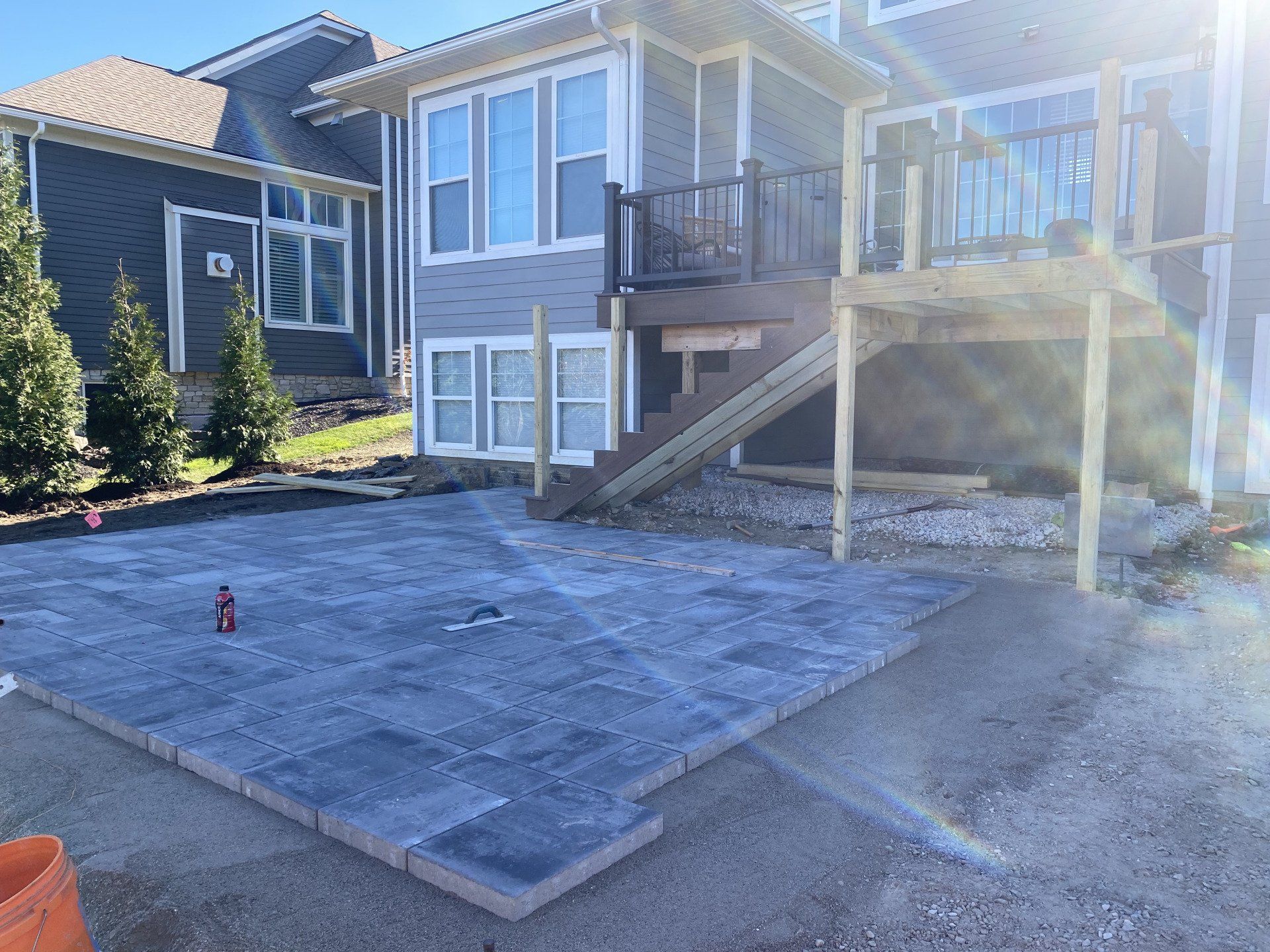 Concrete patio and staircase beside a house, with a sunlit driveway and shrubs nearby