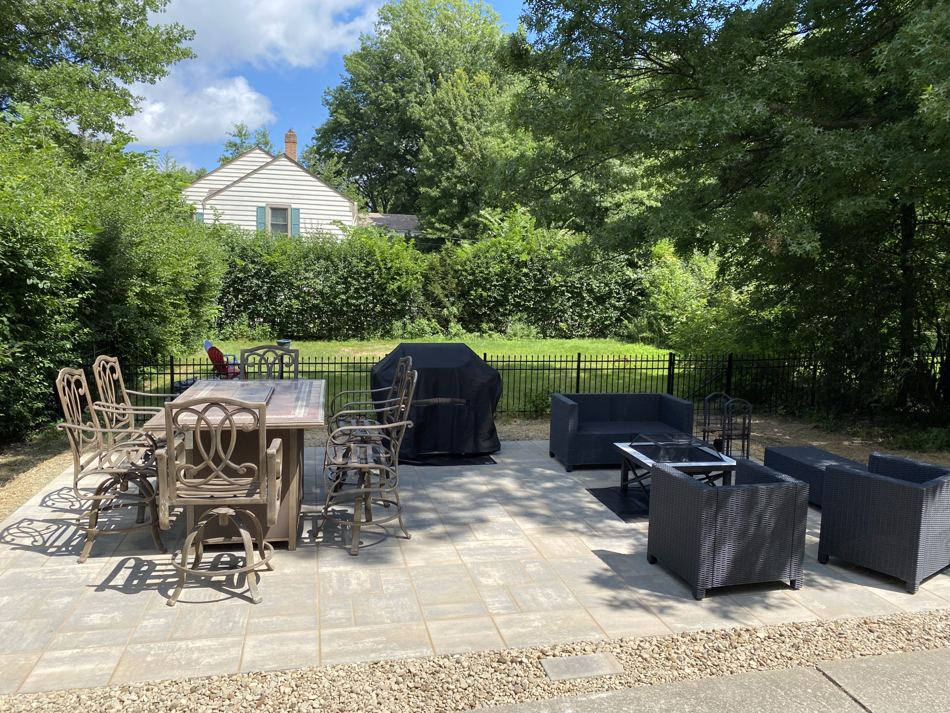Backyard patio with outdoor dining table, grill, and wicker seating amid green hedges and trees