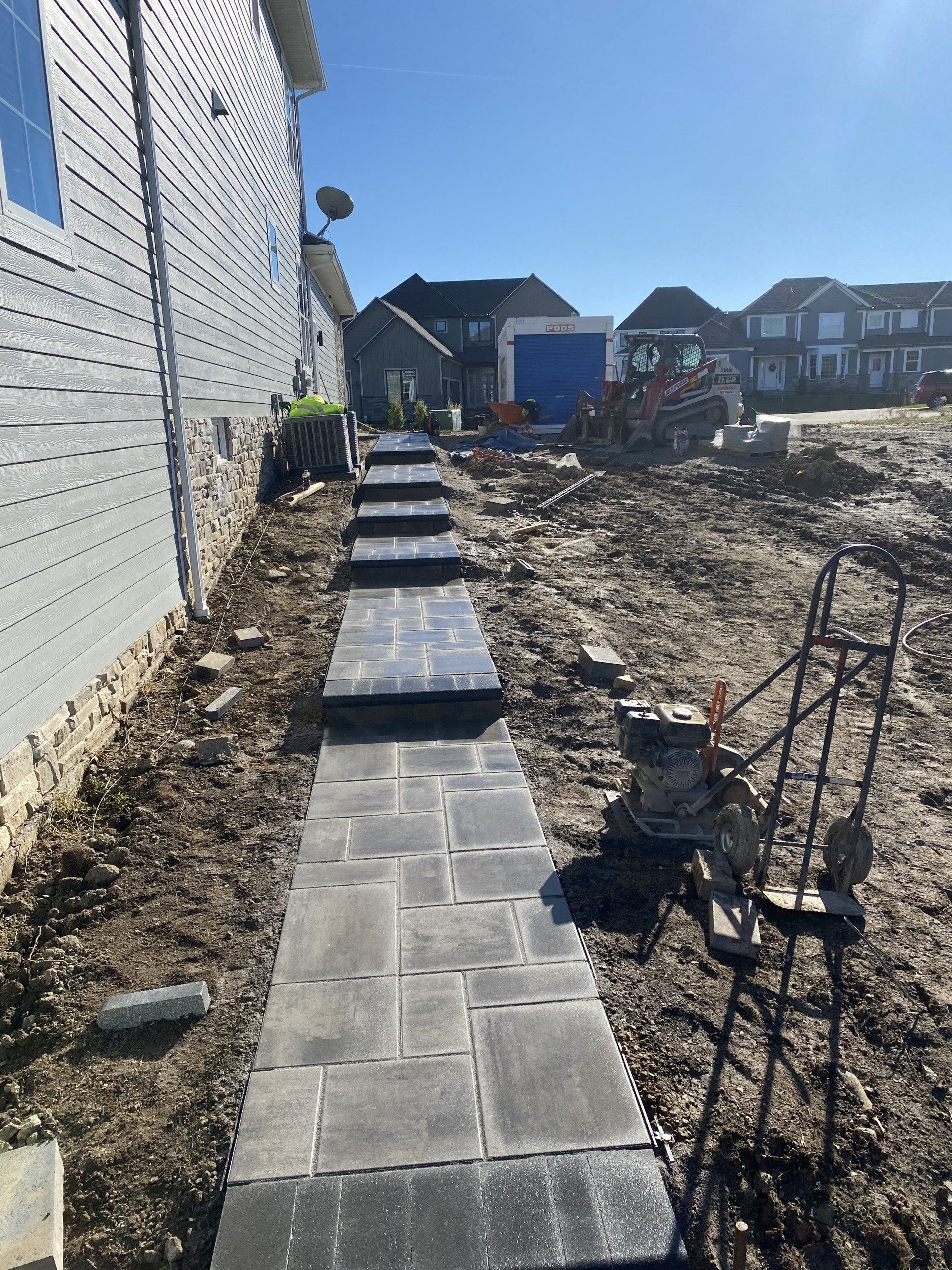 Narrow concrete walkway alongside a house under construction, with dirt yard and steps leading uphill.