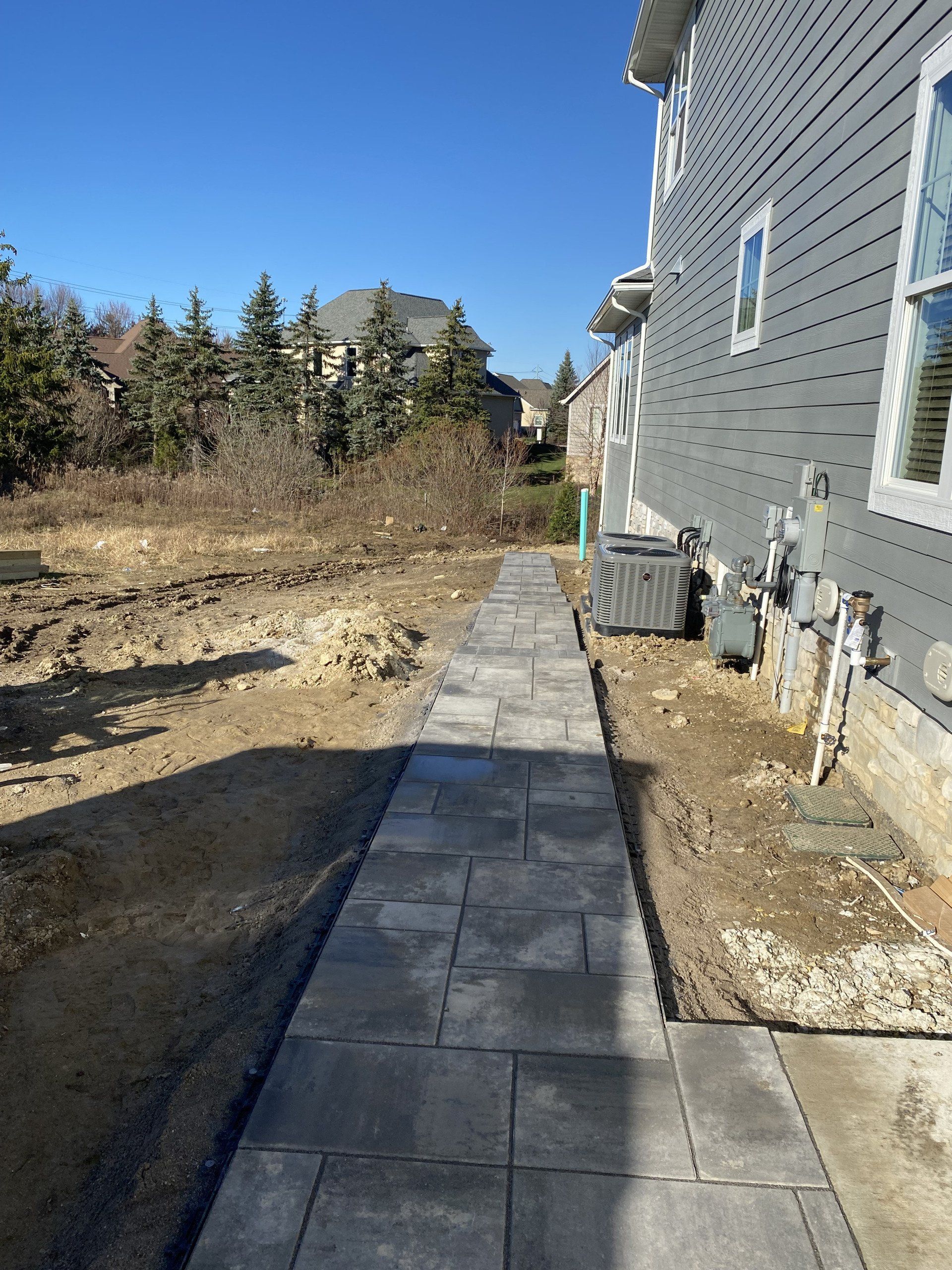 Narrow stone walkway beside a gray house, leading past a dirt yard and shrubs on a sunny day