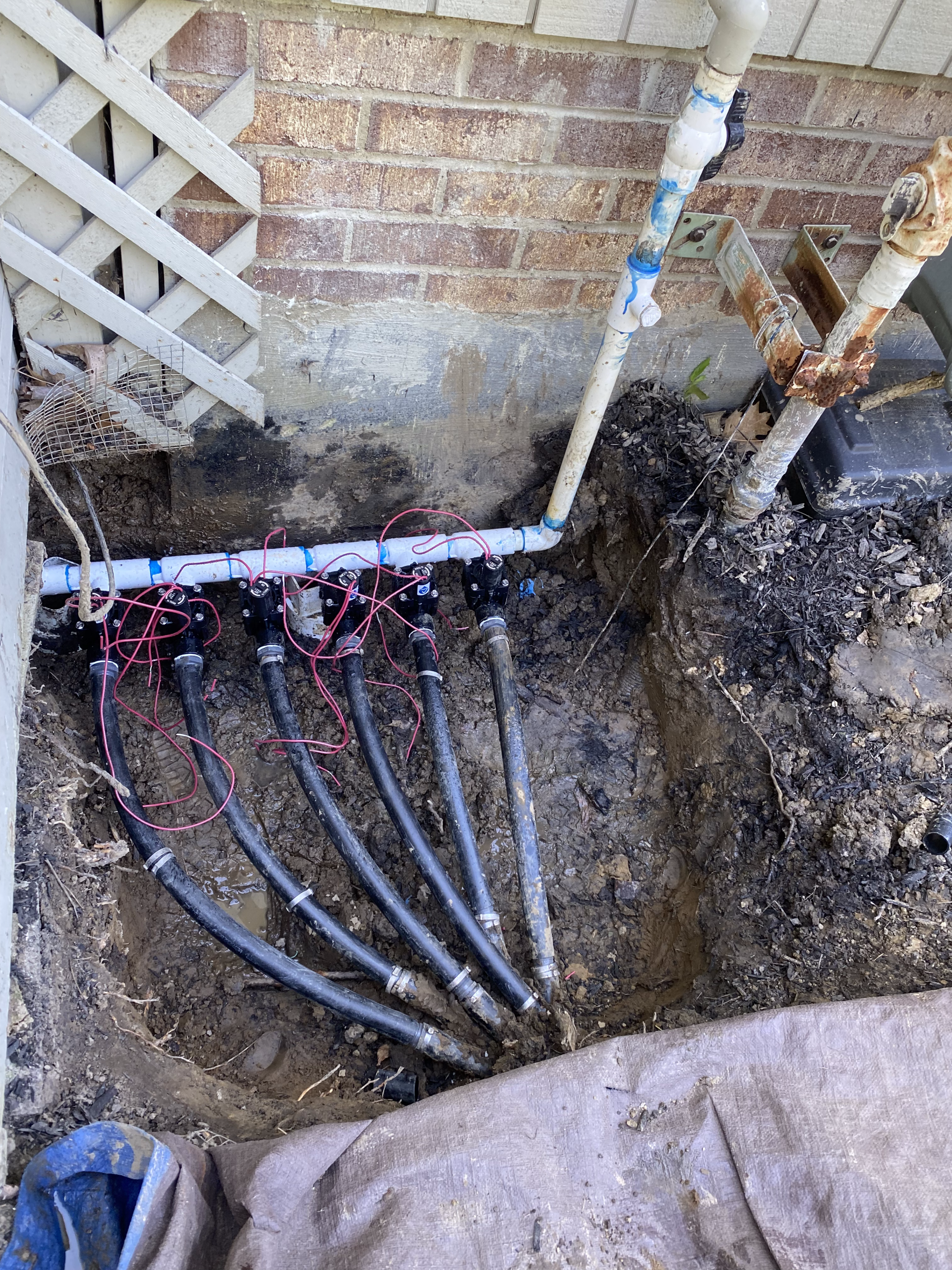 Exposed underground cables and white PVC piping beside a brick foundation wall in a dirt trench.