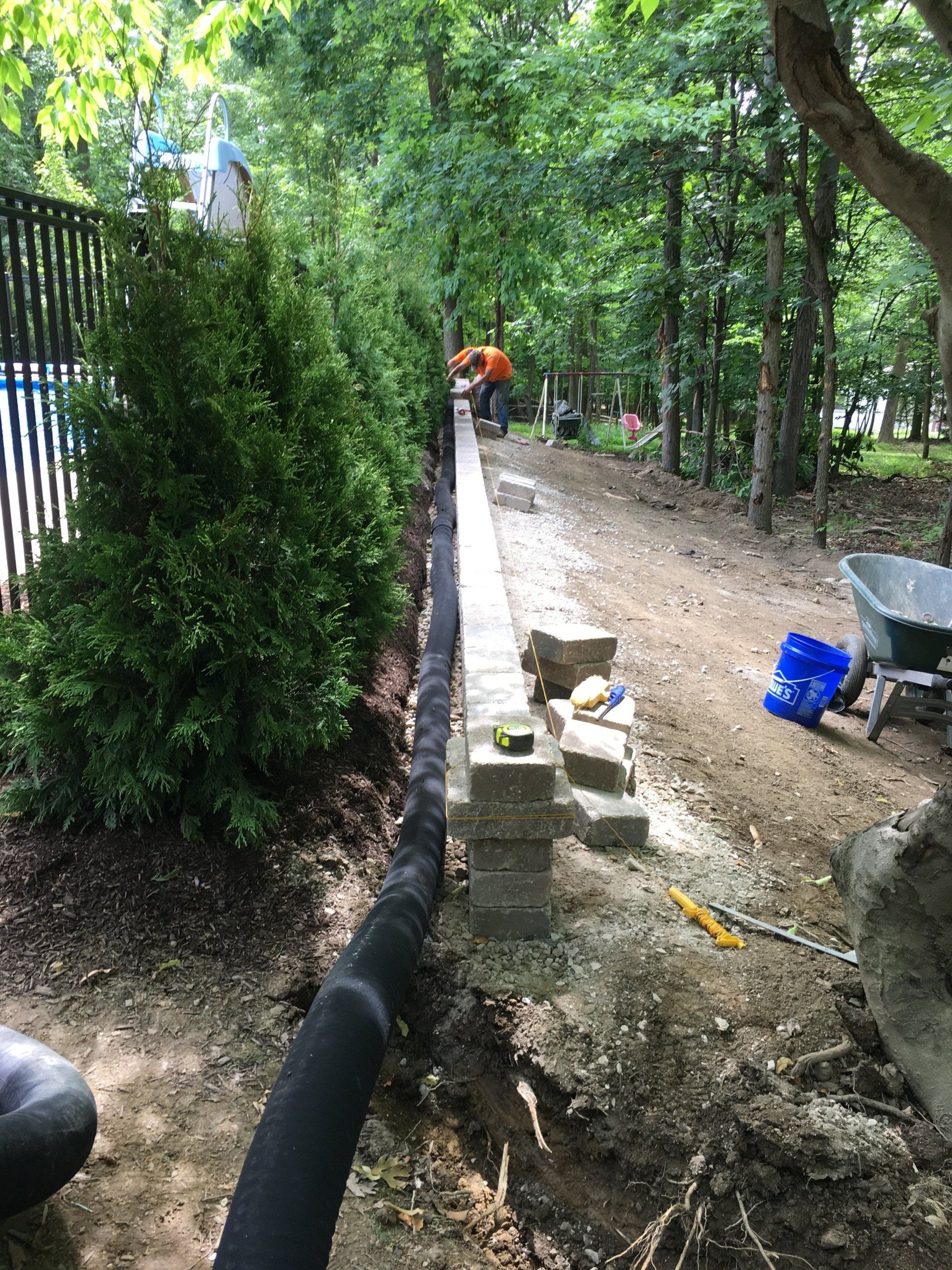 Tree-lined dirt path beside a fence, with a low brick wall and construction tools along the side.