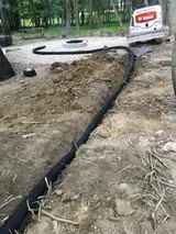 Trenched dirt yard with black drainage pipe, concrete ring, and a white truck near a wooded area