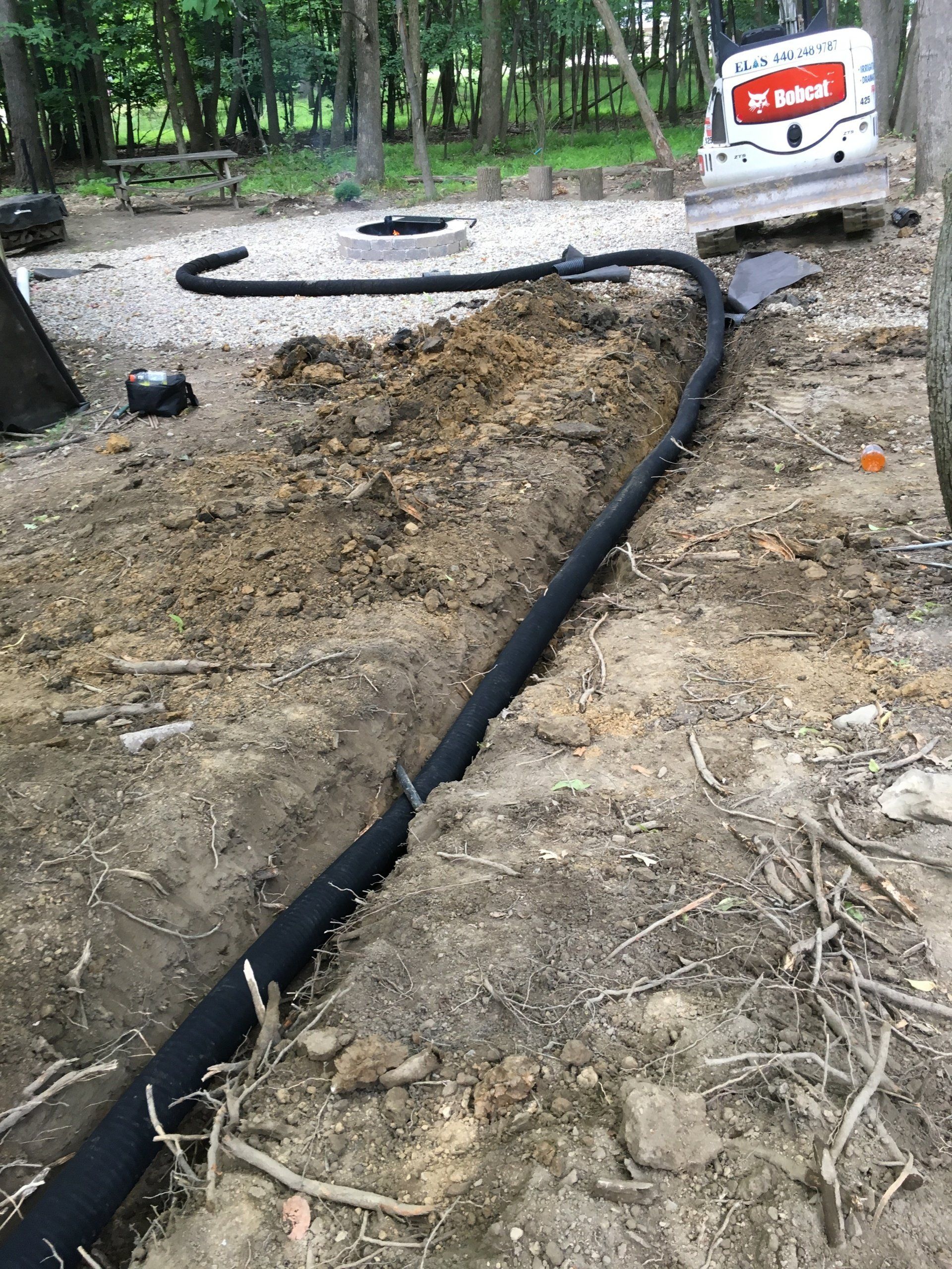 Black drainage pipe in a dug trench at a wooded construction site, with a dump truck in the background