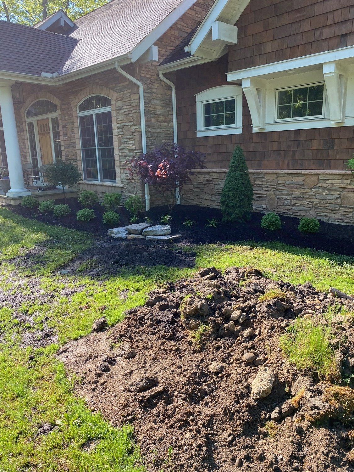 Front yard landscaping with a large pile of dirt, stone house facade, and newly planted shrubs.
