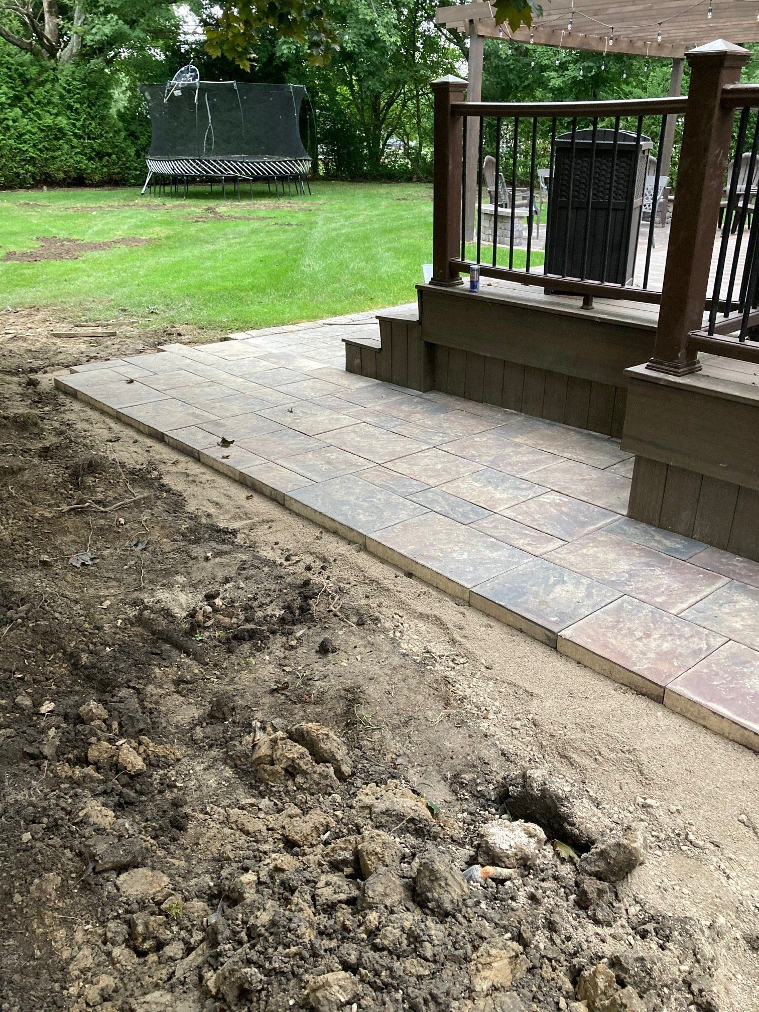 Freshly graded dirt beside a brick patio and wooden porch in a green backyard