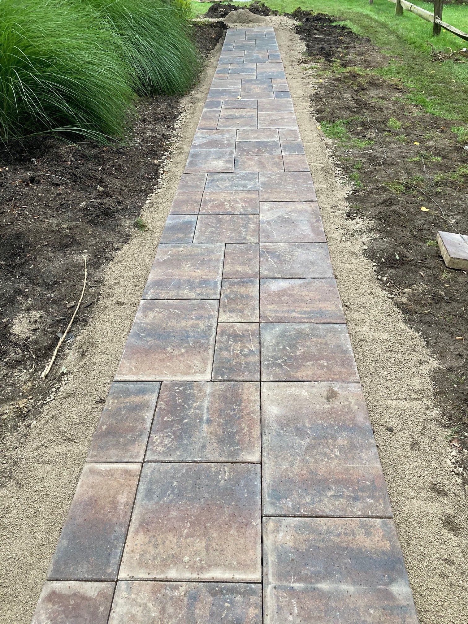 Narrow stone garden walkway bordered by soil and green plants.