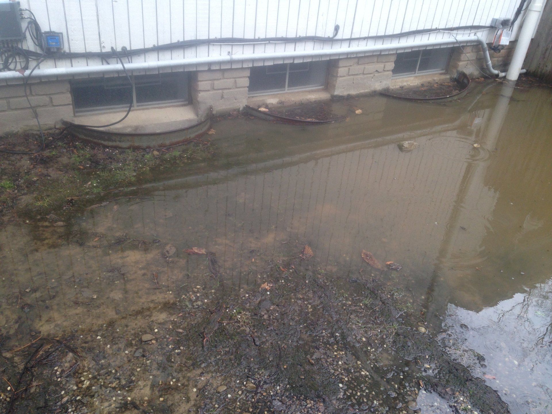 Mud and standing water beside a white building wall and fence after rain.