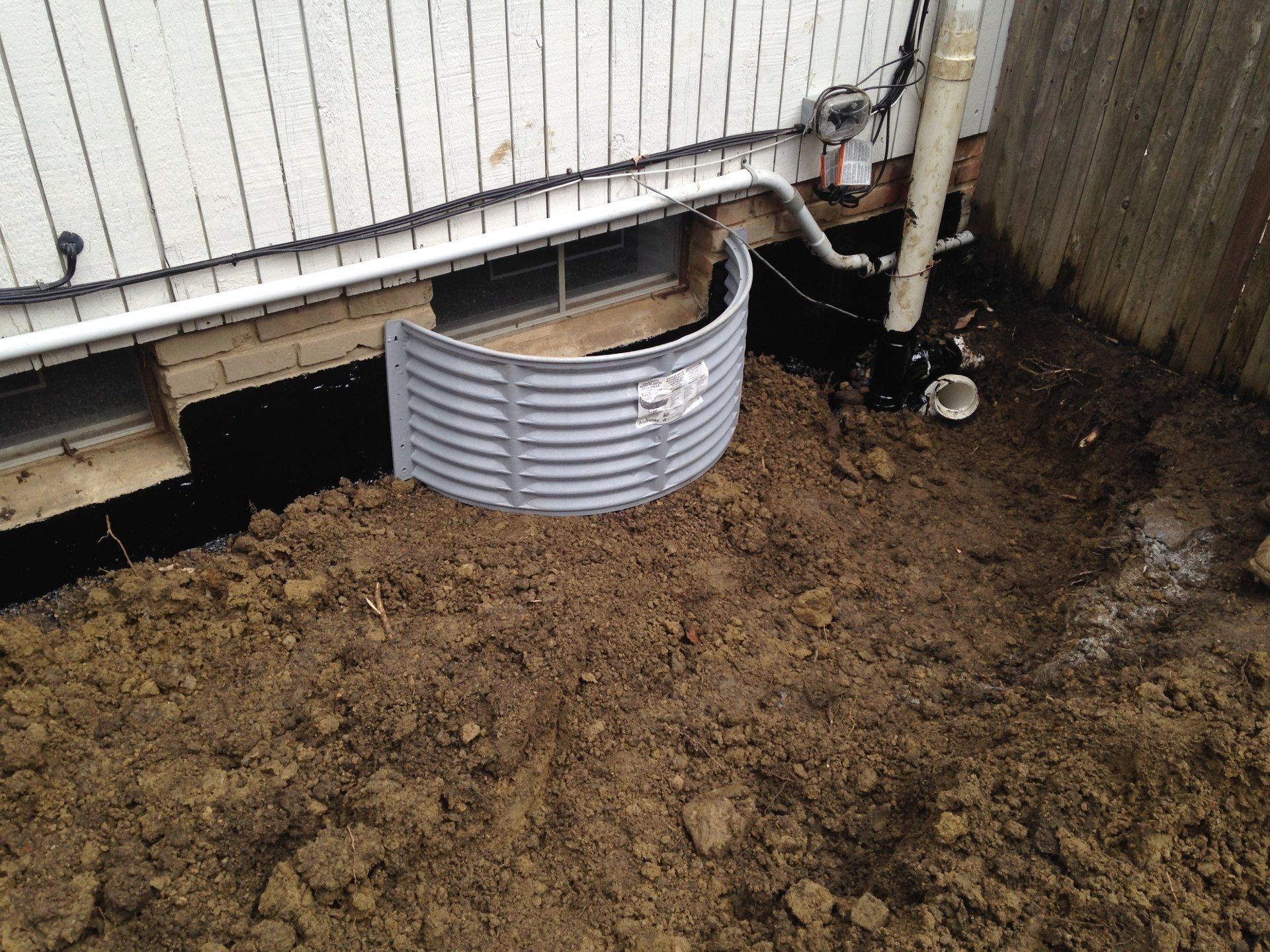 Muddy backyard beside a white house wall with a small gray corrugated barrier and exposed foundation area