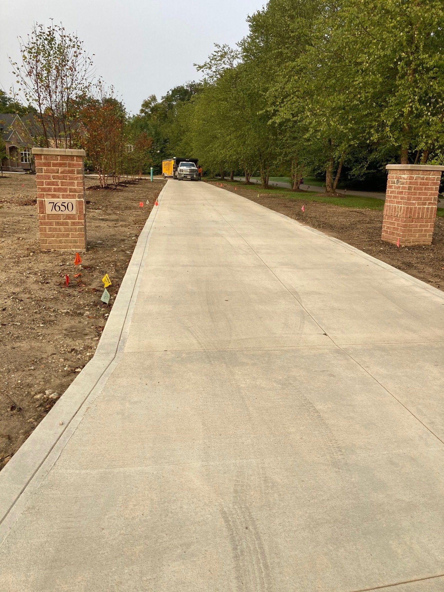 Concrete driveway lined with trees and brick pillars in a rural setting