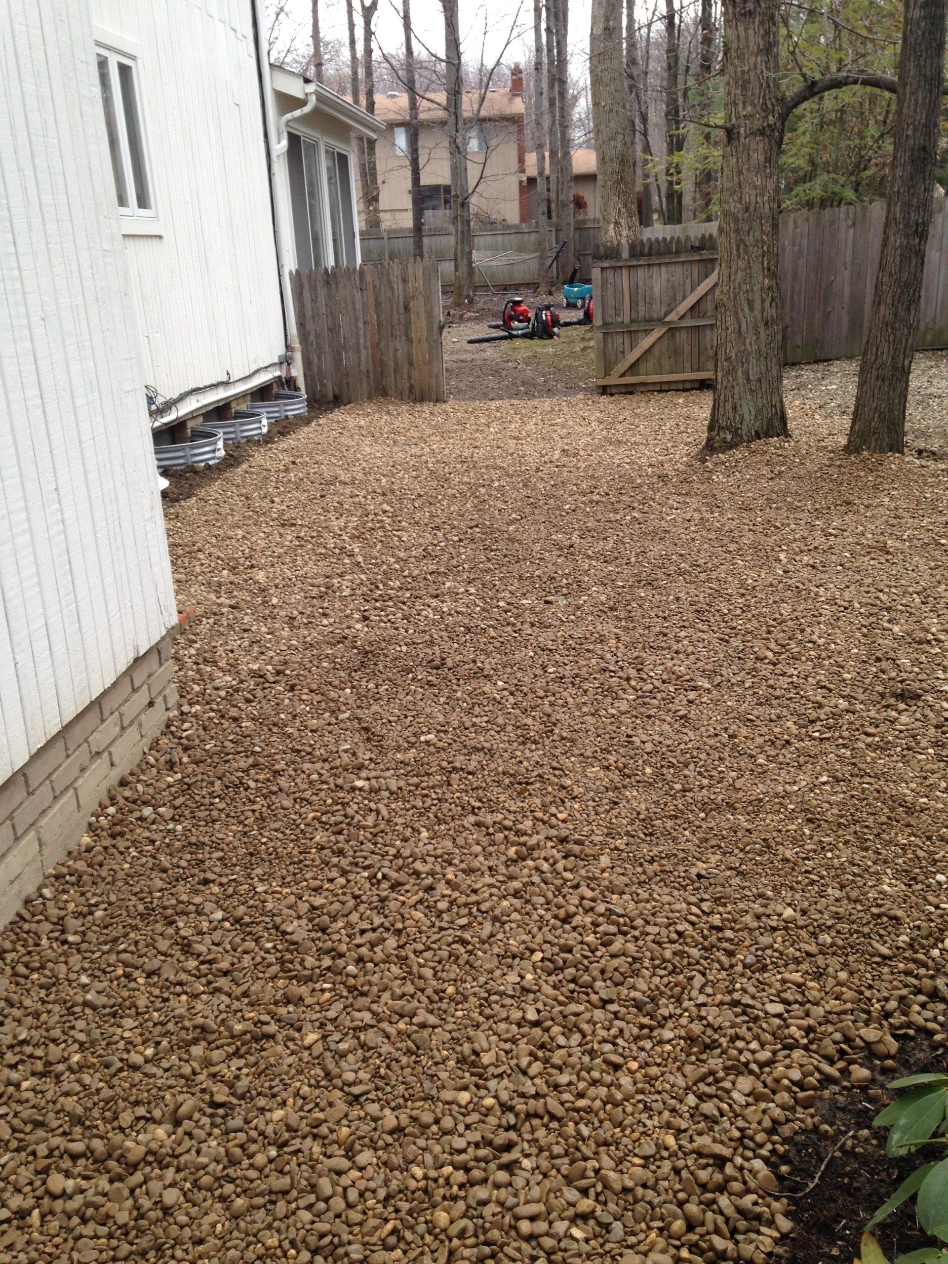 Backyard covered in tan gravel beside a white house, with trees and a wooden gate in the distance.