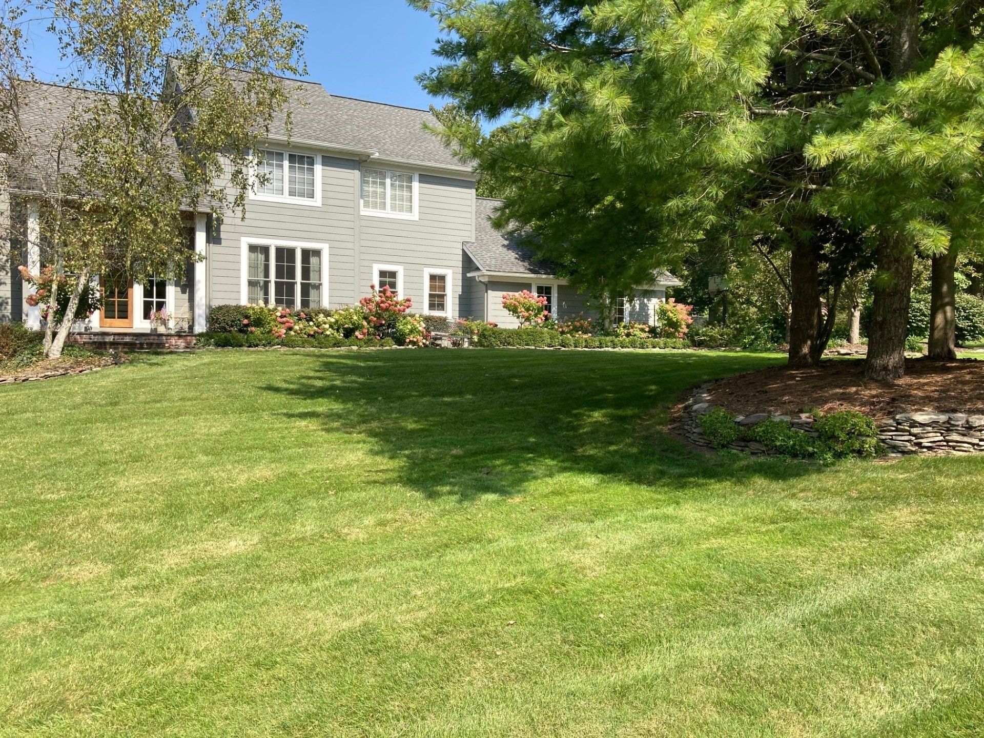 Gray house with white trim beside a large green lawn and trees on a sunny day