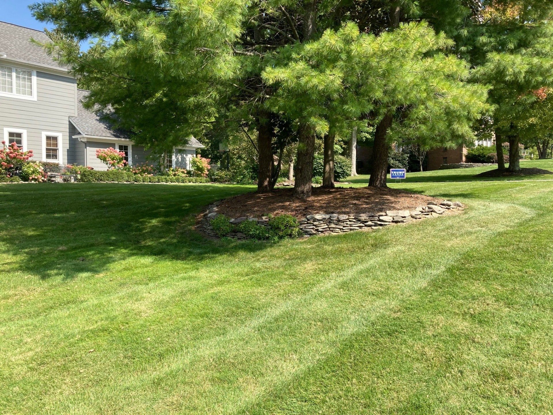 Tree-shaded lawn beside a gray house with a stone-mulched bed and bright green grass