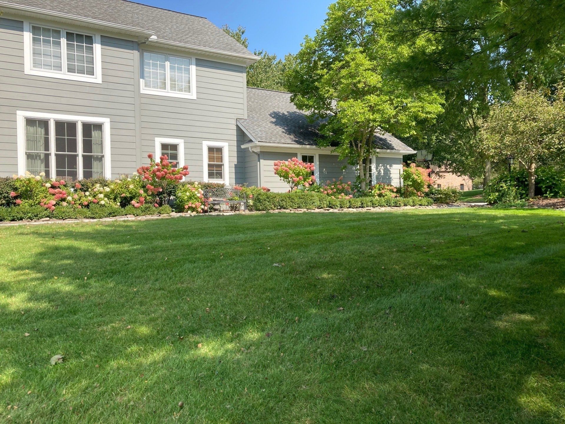 Gray house with white trim beside a green lawn and flower bed under trees