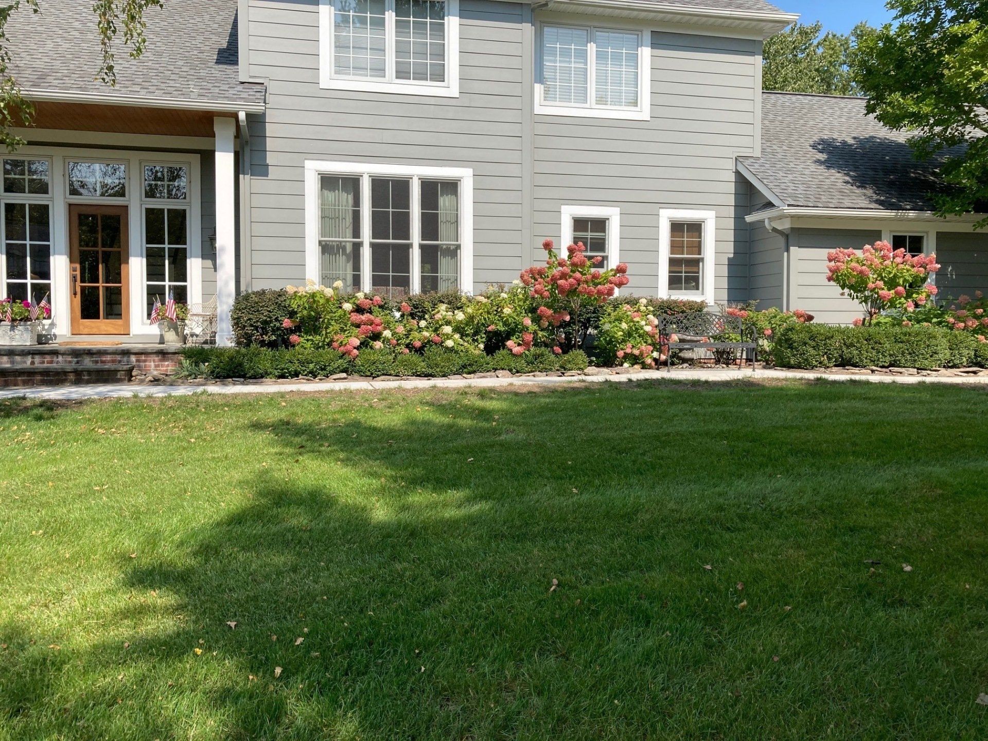 Gray house with white trim, flower beds, and a green front lawn under bright sunlight