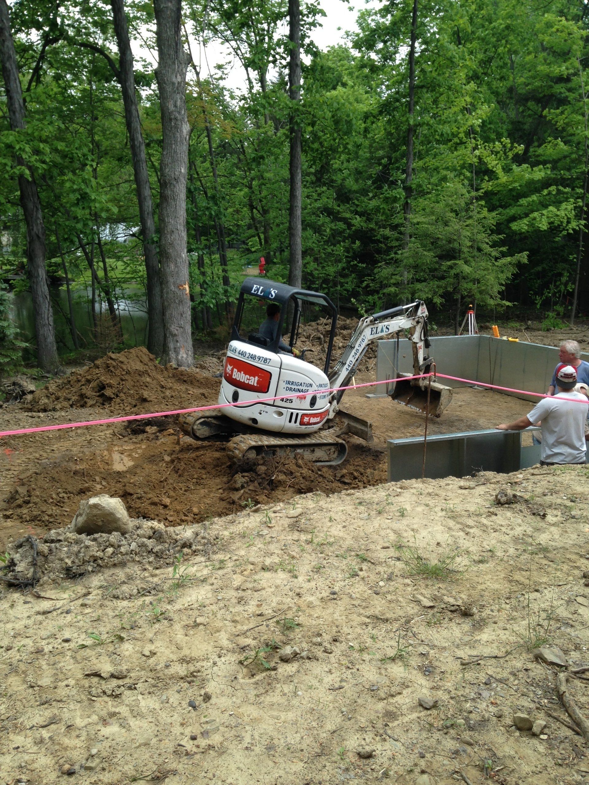 Small excavator digging a muddy trench beside a wooded creek, with a worker nearby.