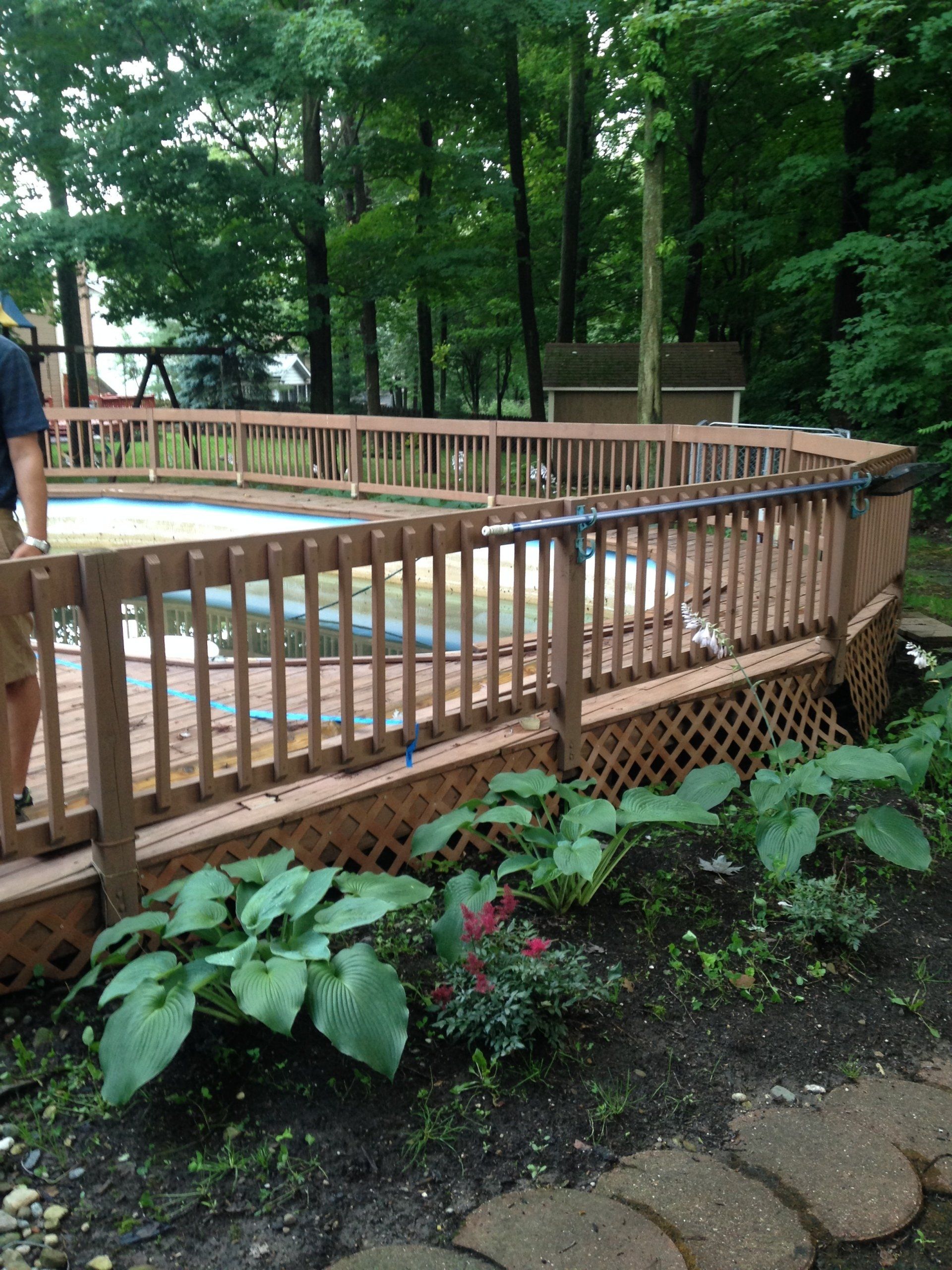 Wooden deck surrounding a small above-ground pool in a wooded backyard with flowers and stone edging.