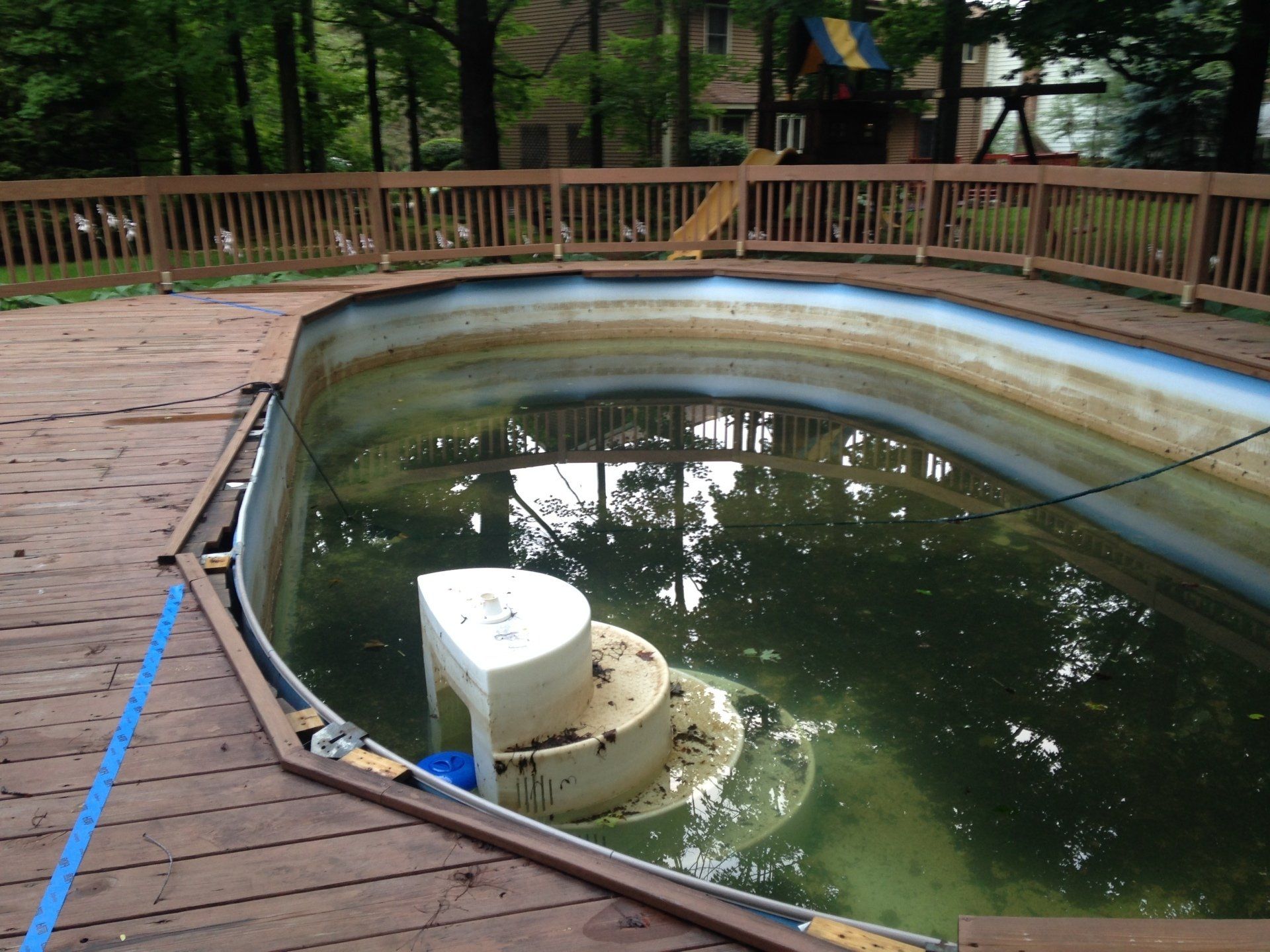 Empty backyard pool with green water and a wooden deck railing, surrounded by trees