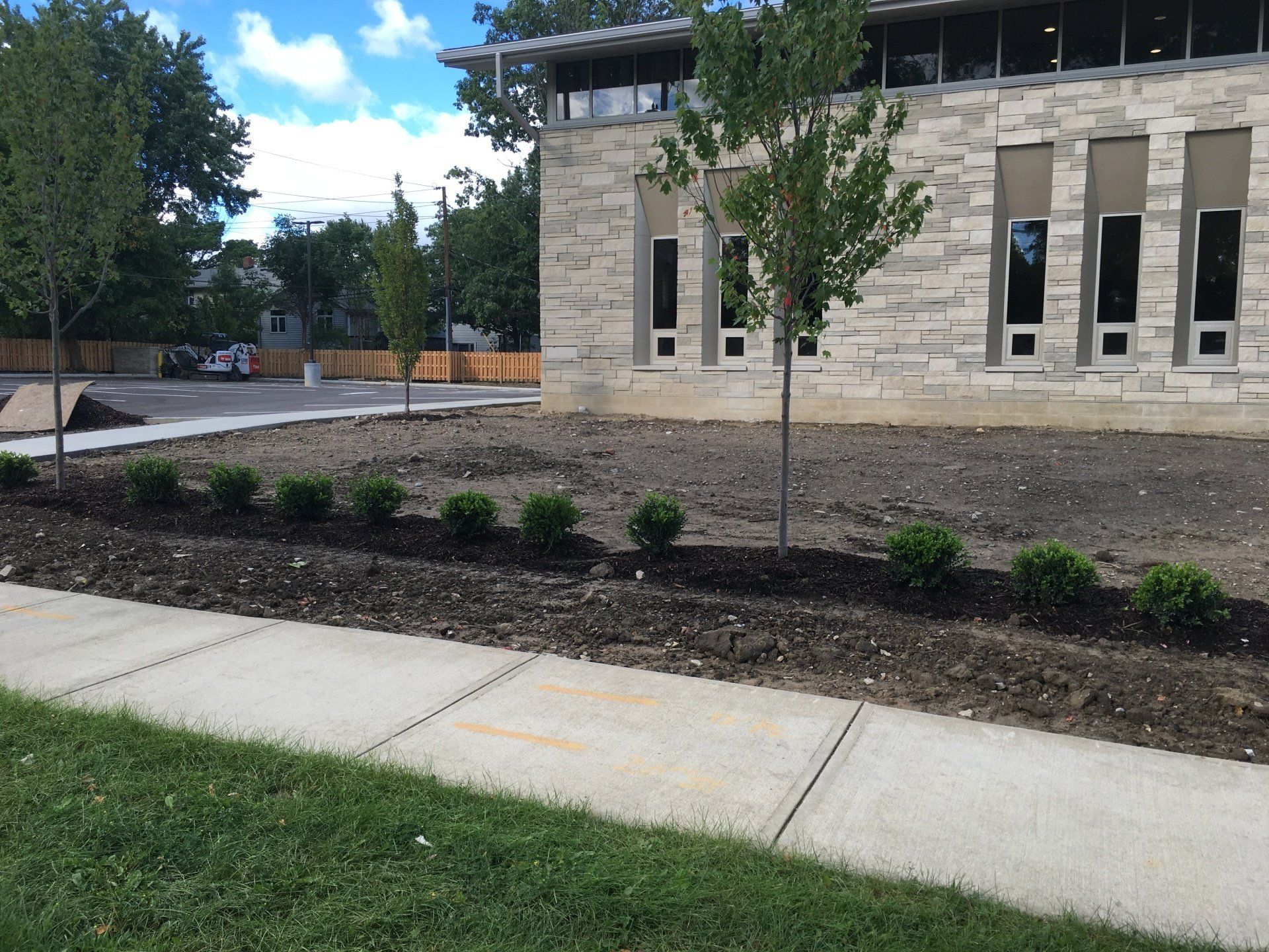 Stone building beside a sidewalk and landscaped mulch beds with small shrubs and a tree