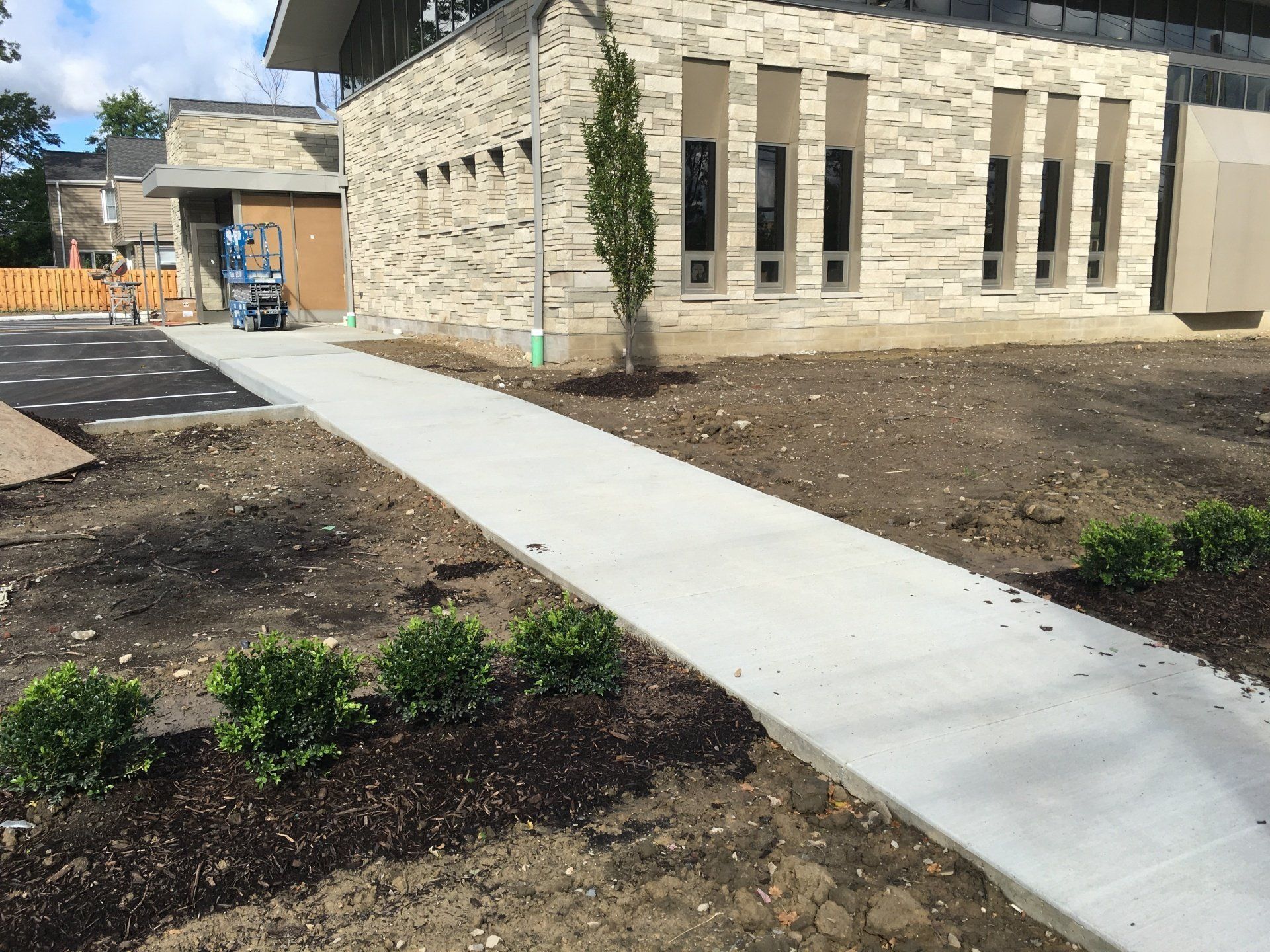 Concrete sidewalk beside a beige stone building with newly landscaped mulch beds and small shrubs