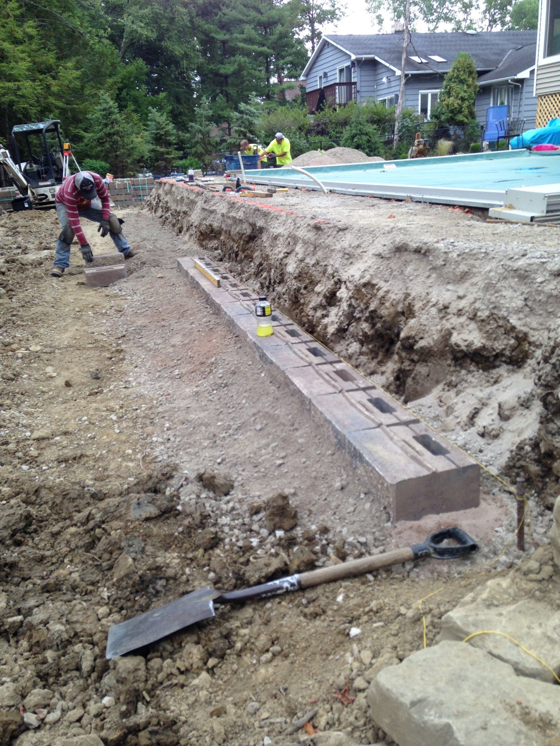 Construction worker grading a dirt trench beside a low brick wall at a house site