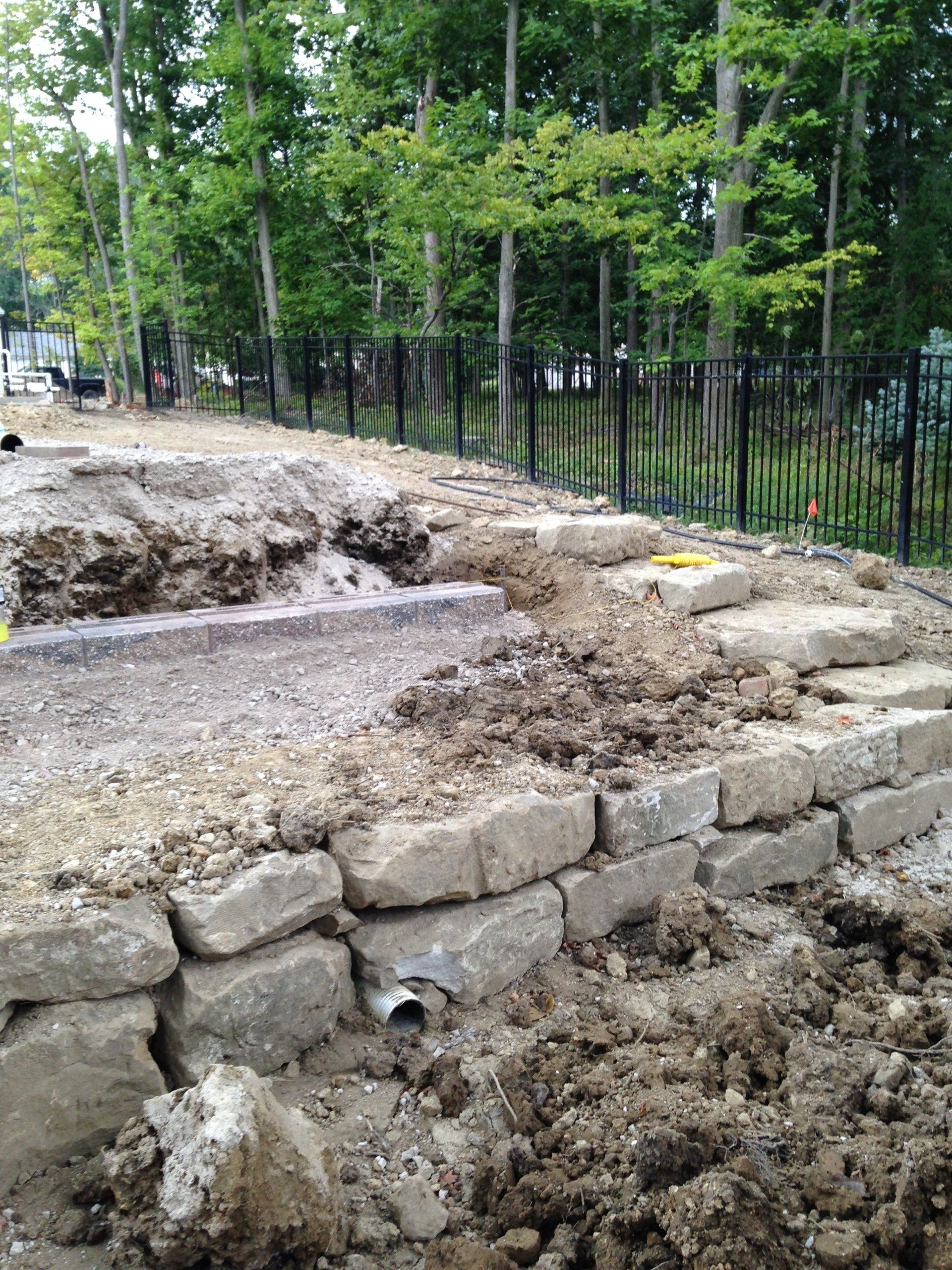 Stone retaining wall under construction beside a black metal fence and trees