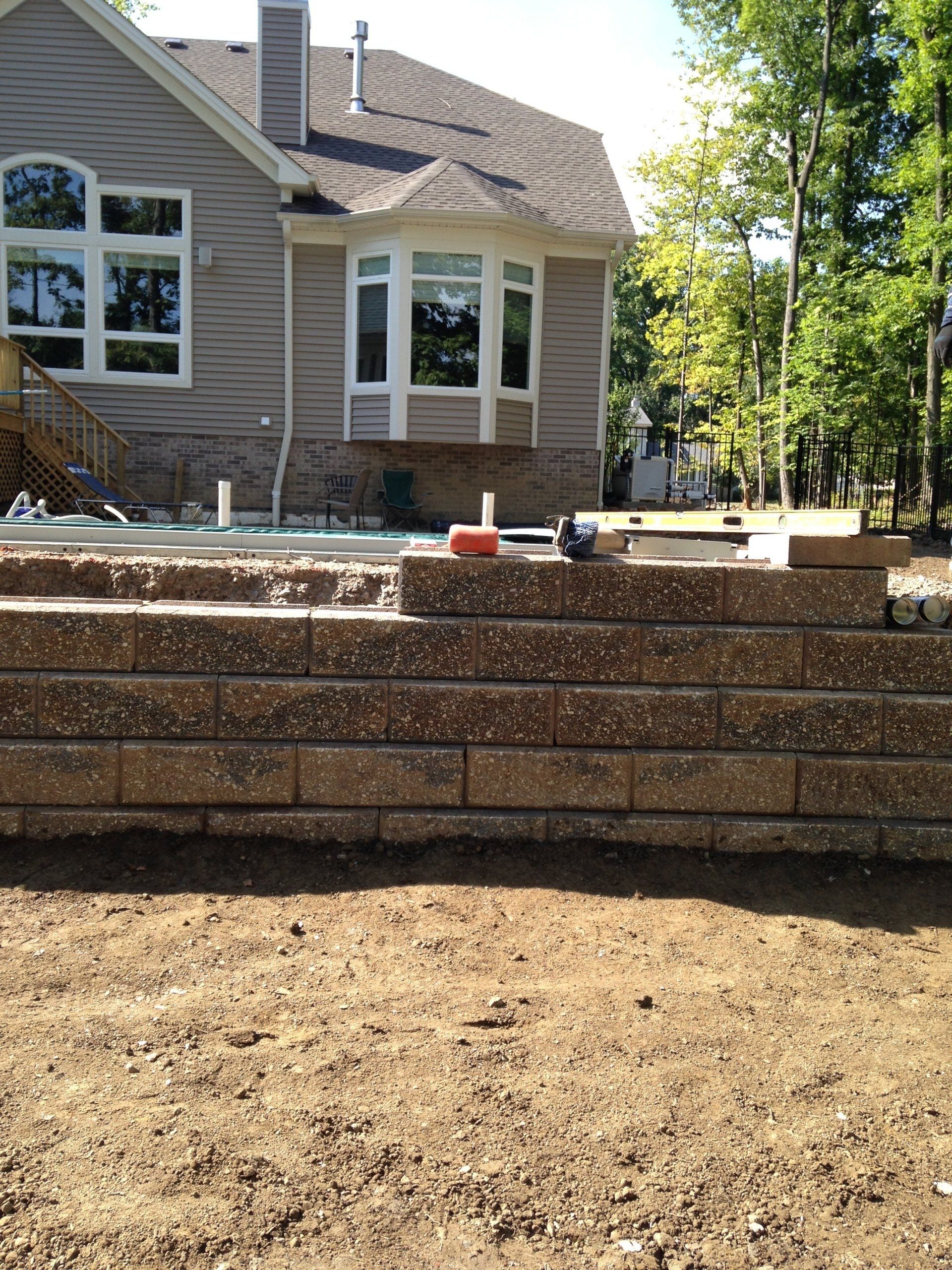 Backyard with terraced retaining wall, bare dirt foreground, and house with large windows in the background.