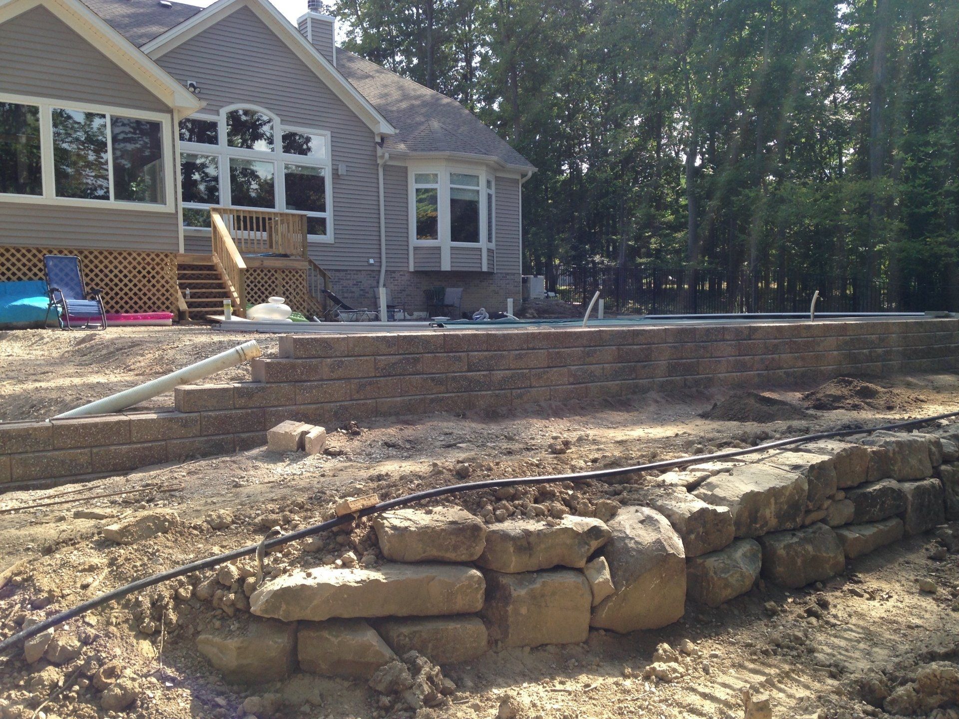 Stone retaining wall in a backyard beside a house with a patio and wooded trees behind it