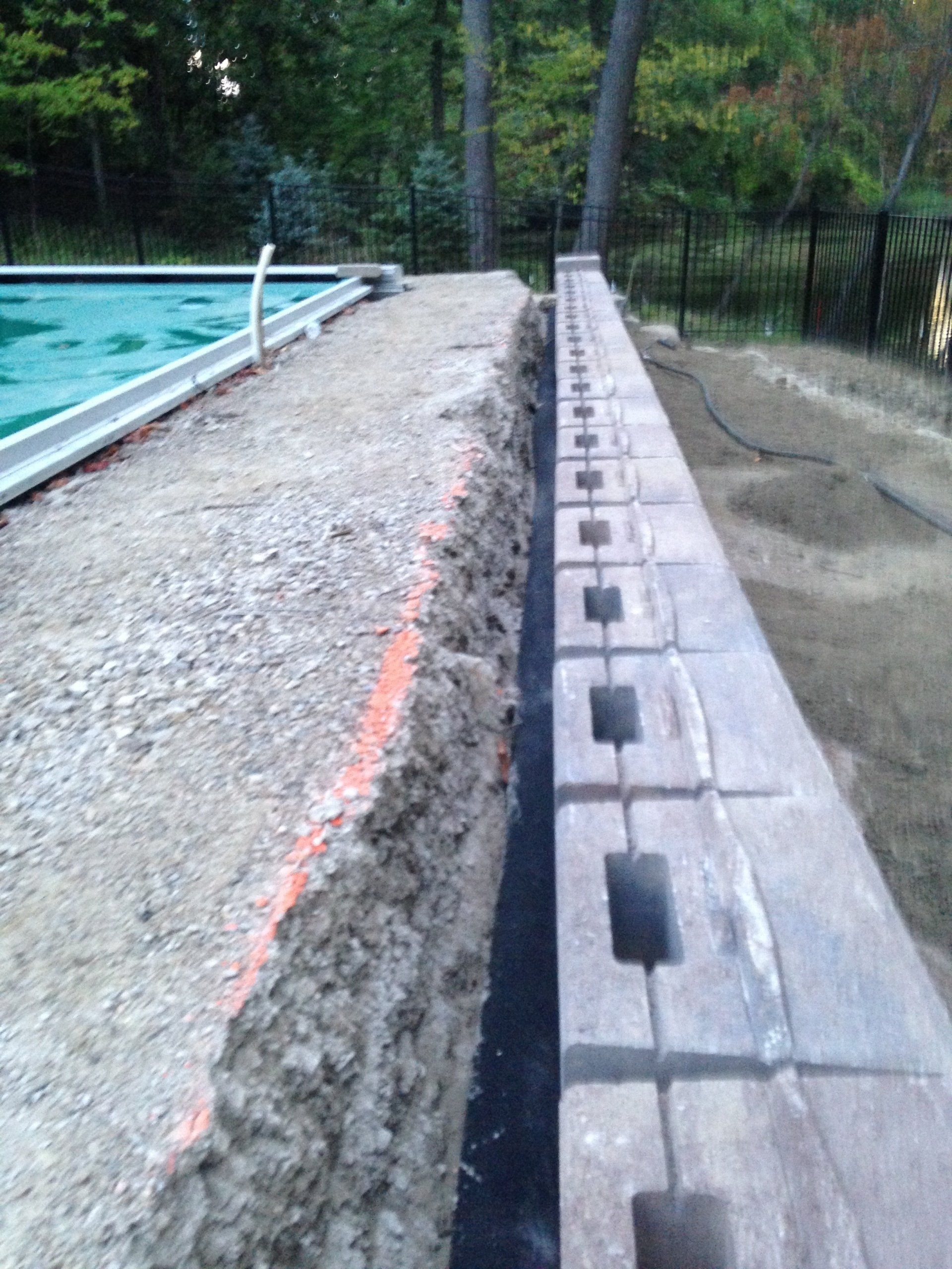 Concrete pool edge beside a gravel path and retaining wall, with trees and a fence in the background