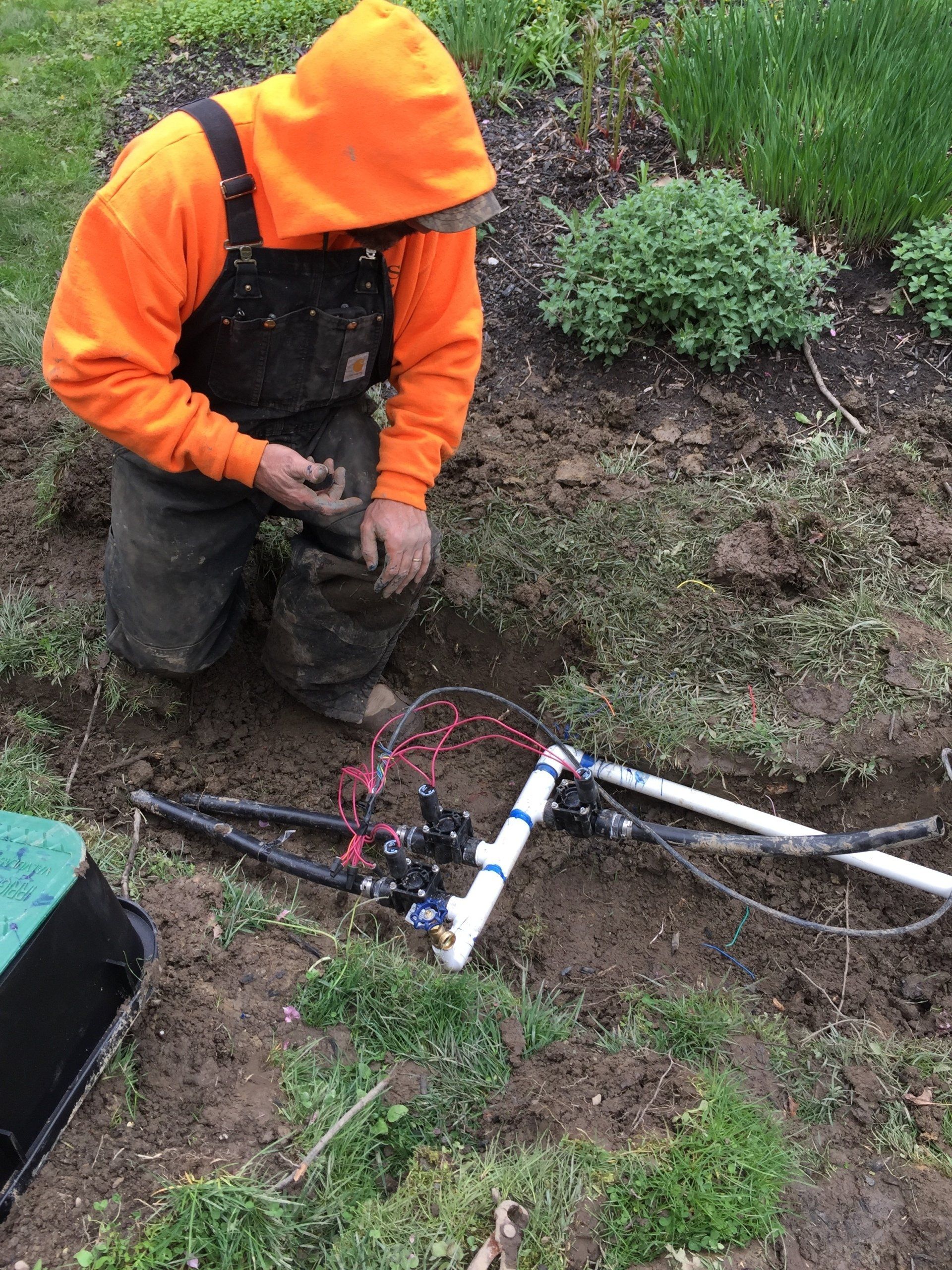 Worker in orange rain gear kneels by a trench, adjusting a cable or pipe in wet ground.