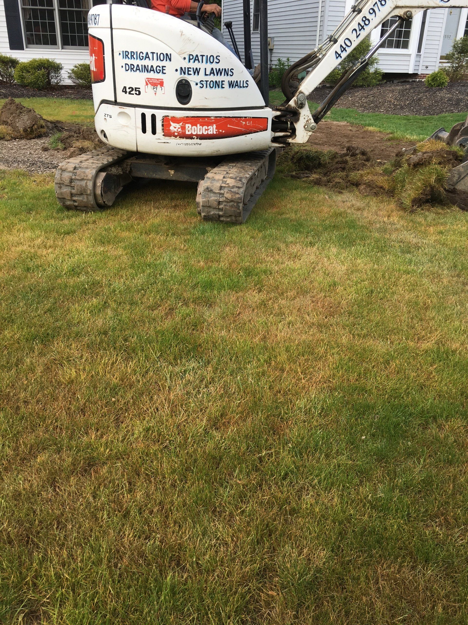 Small excavator on a grassy lawn near a building, with muddy tracks and a white cab.