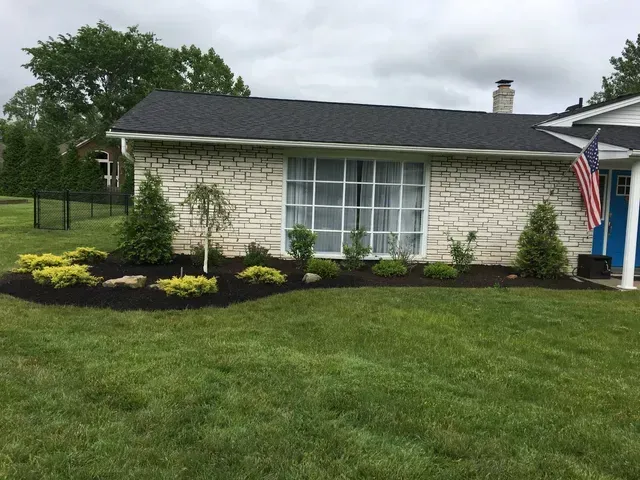 Small stone house with white windows, dark roof, landscaped shrubs, green lawn, and an American flag by the porch