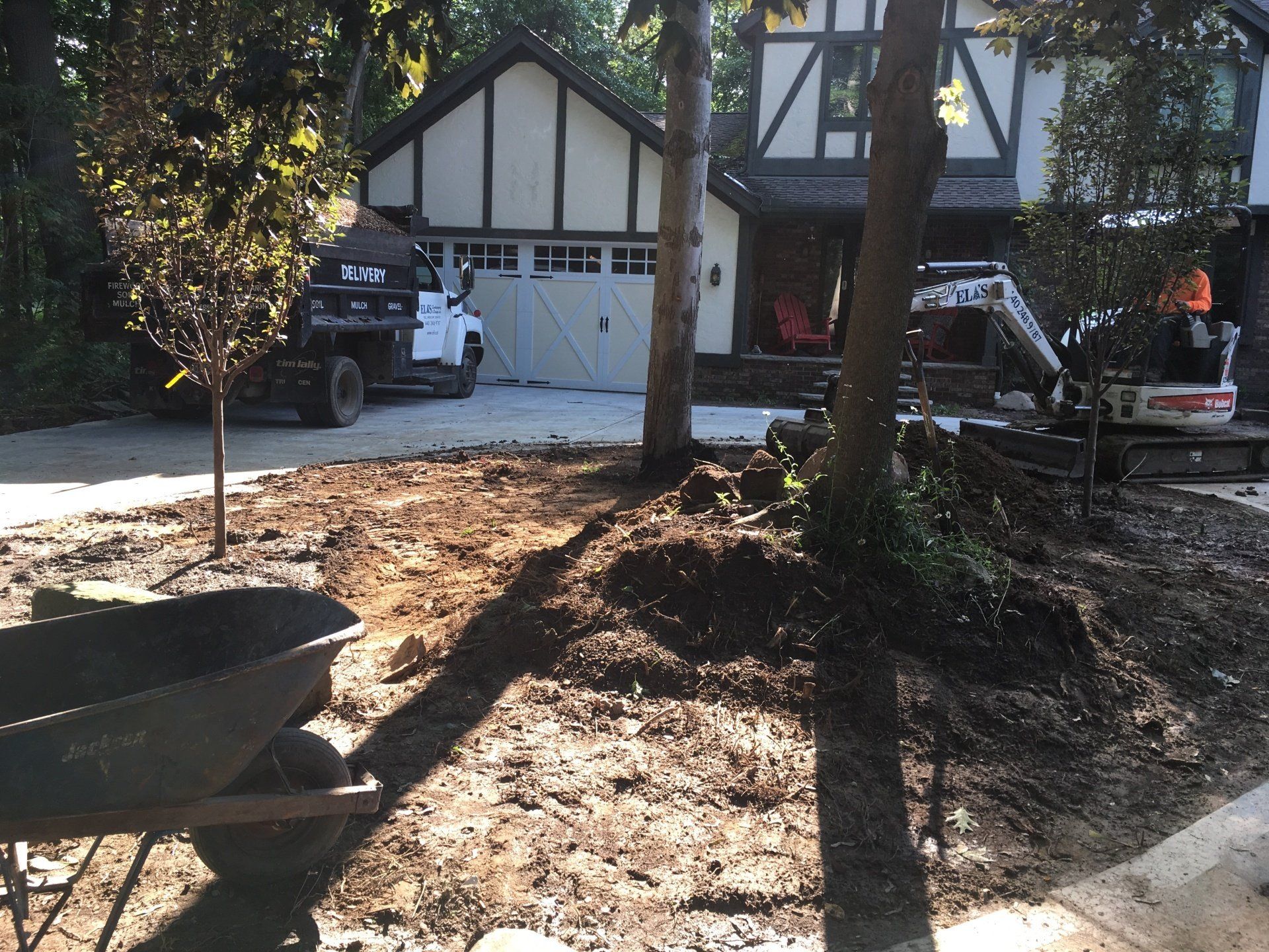 Front yard with a truck, wheelbarrow, and house under trees, showing ongoing landscaping work.