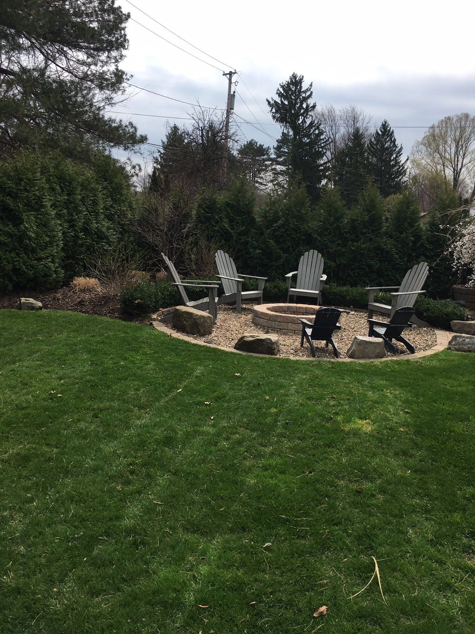 Backyard patio with white chairs around a fire pit, surrounded by green lawn and trees under a cloudy sky