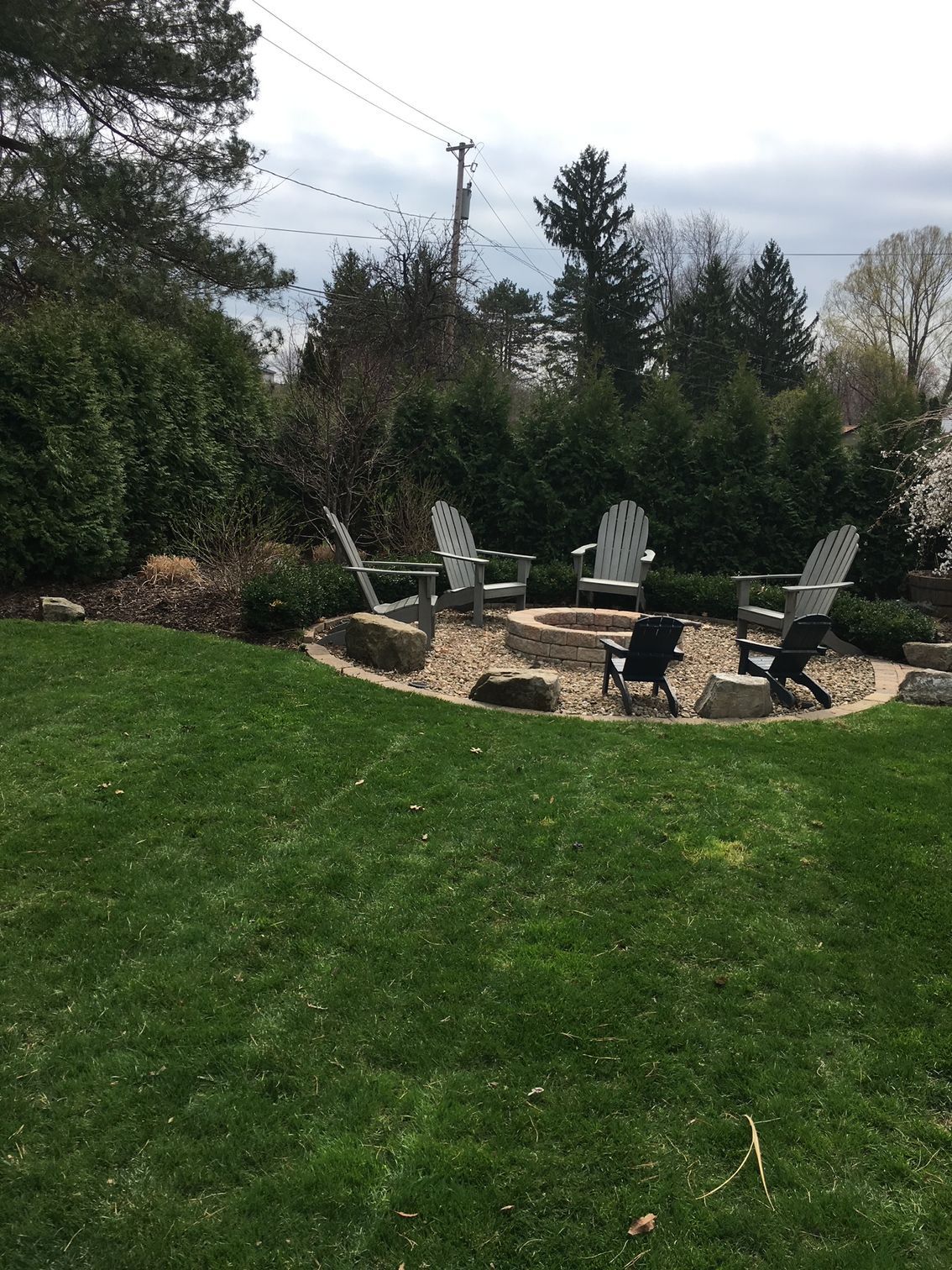 Backyard patio with four white chairs around a stone fire pit, surrounded by grass and trees.