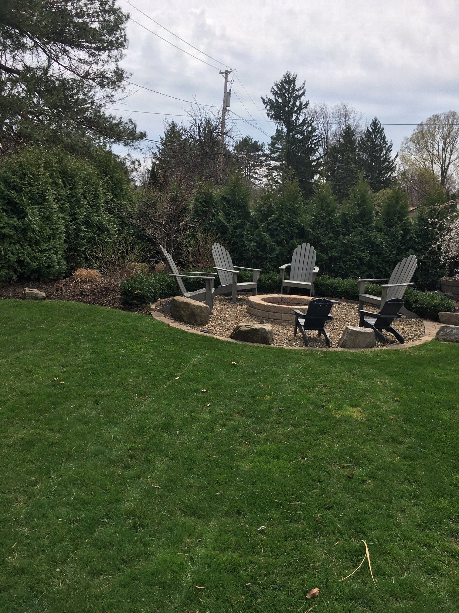 Backyard patio with chairs around a fire pit, surrounded by green lawn and trees