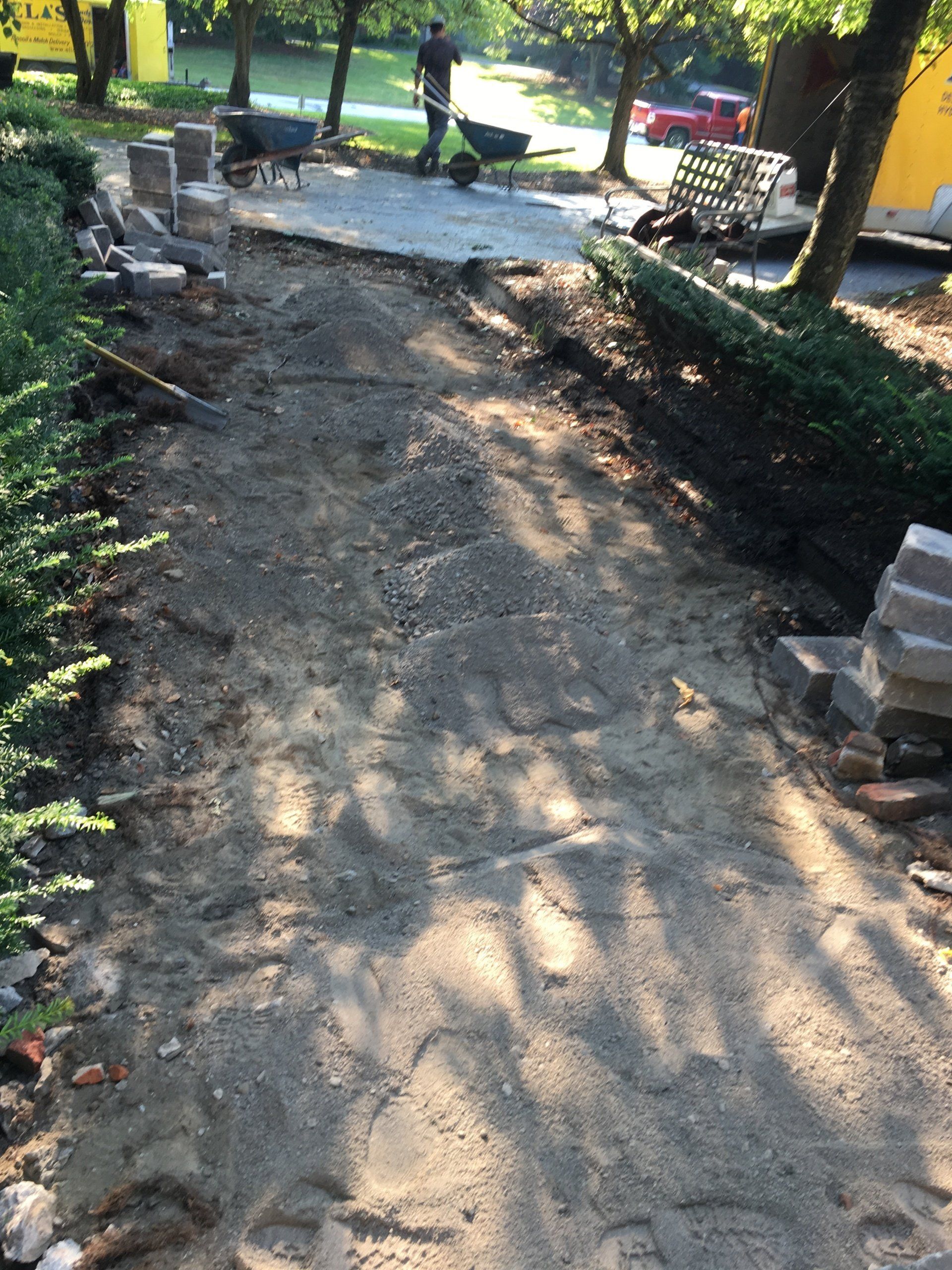 Stone walkway under construction in a park, with workers and stacked pavers near trees and shrubs.