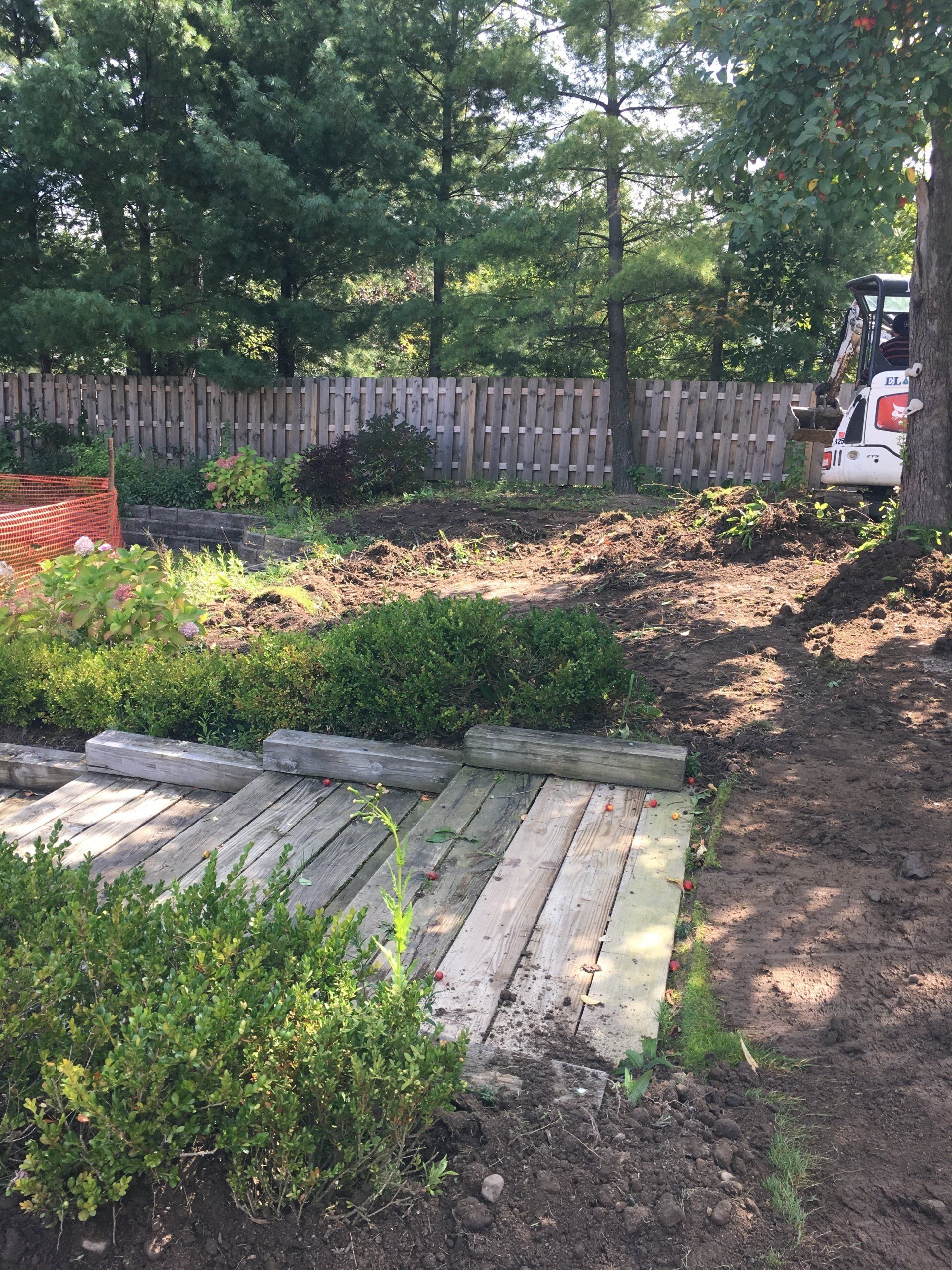Backyard garden with wooden steps, dirt path, shrubs, and a white vehicle by a fence