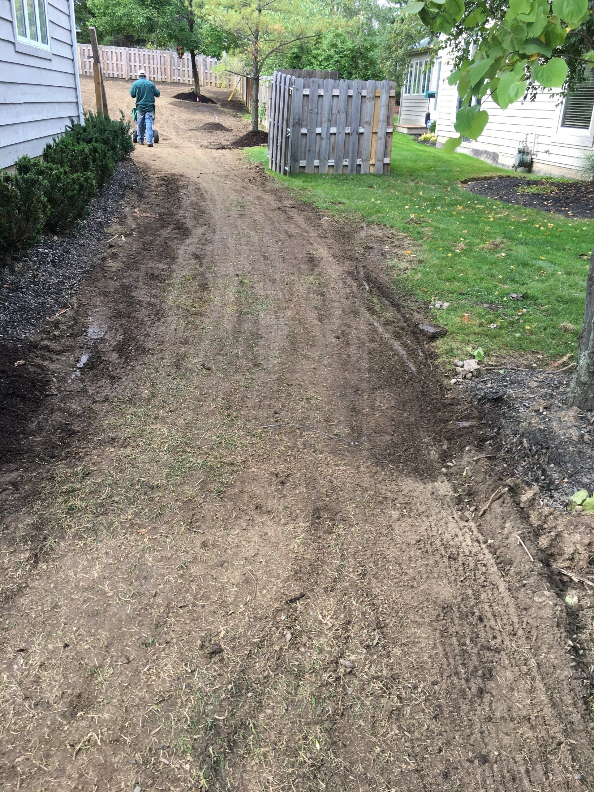 Dirt path between houses and fence, with a person in the distance and a hose or trench along the ground