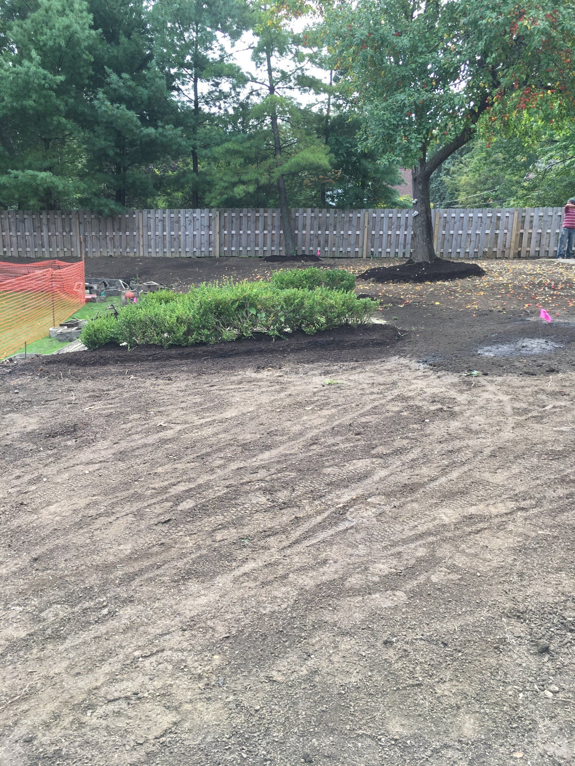 Gravel yard with a small garden bed, wooden fence, orange safety netting, and trees in the background