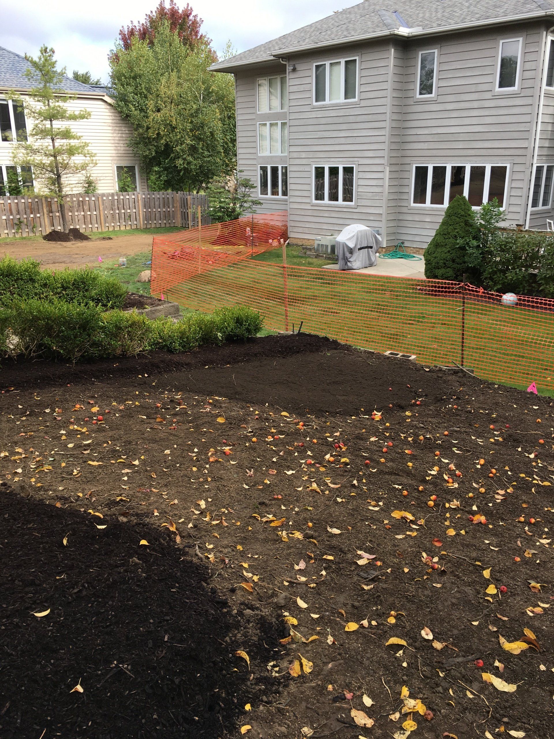 Backyard construction site with dark soil, orange safety fencing, and a gray apartment building in the background