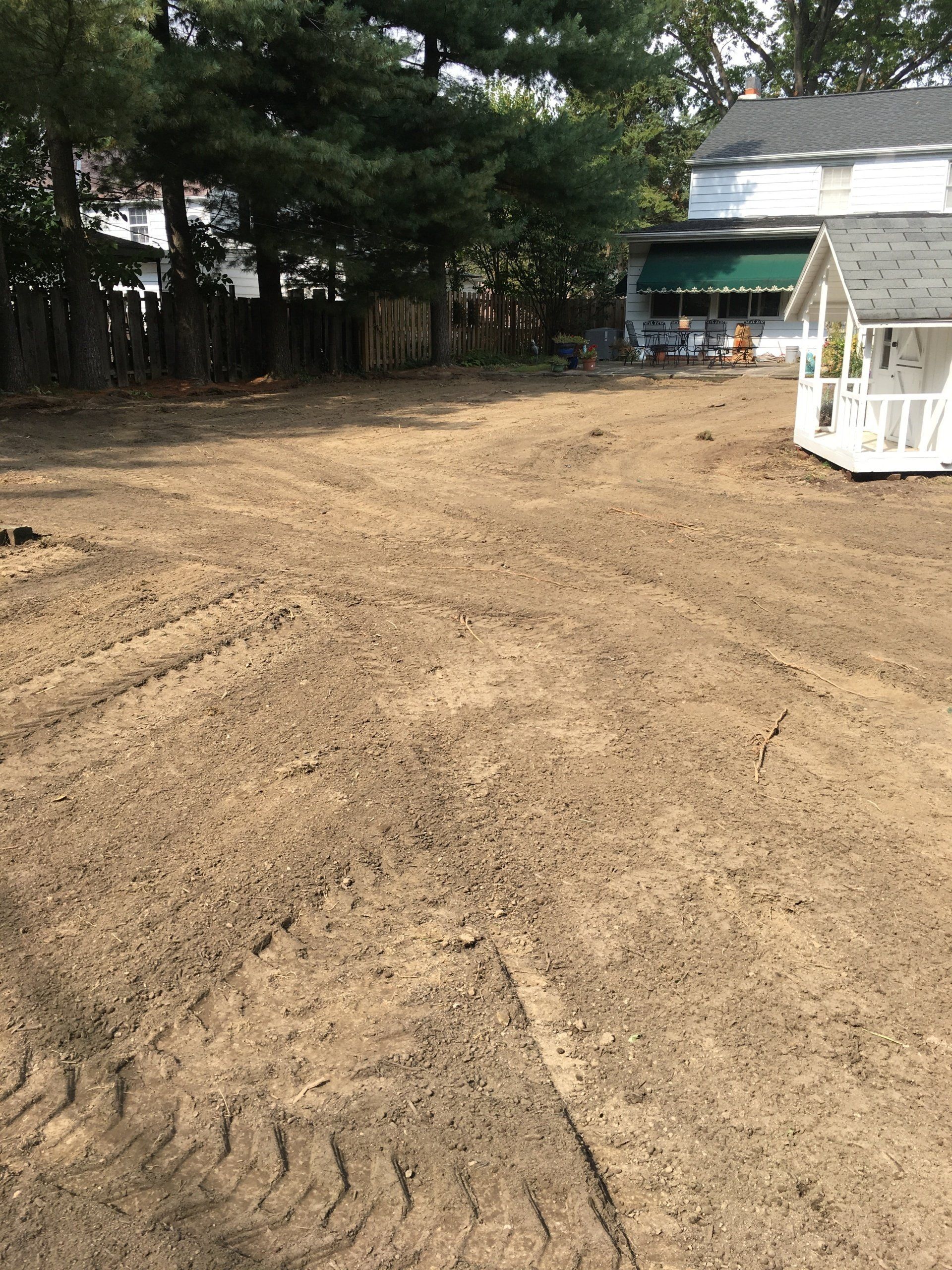 Bare yard with fresh dirt, trees along the left, and a small white playhouse near a house on the right