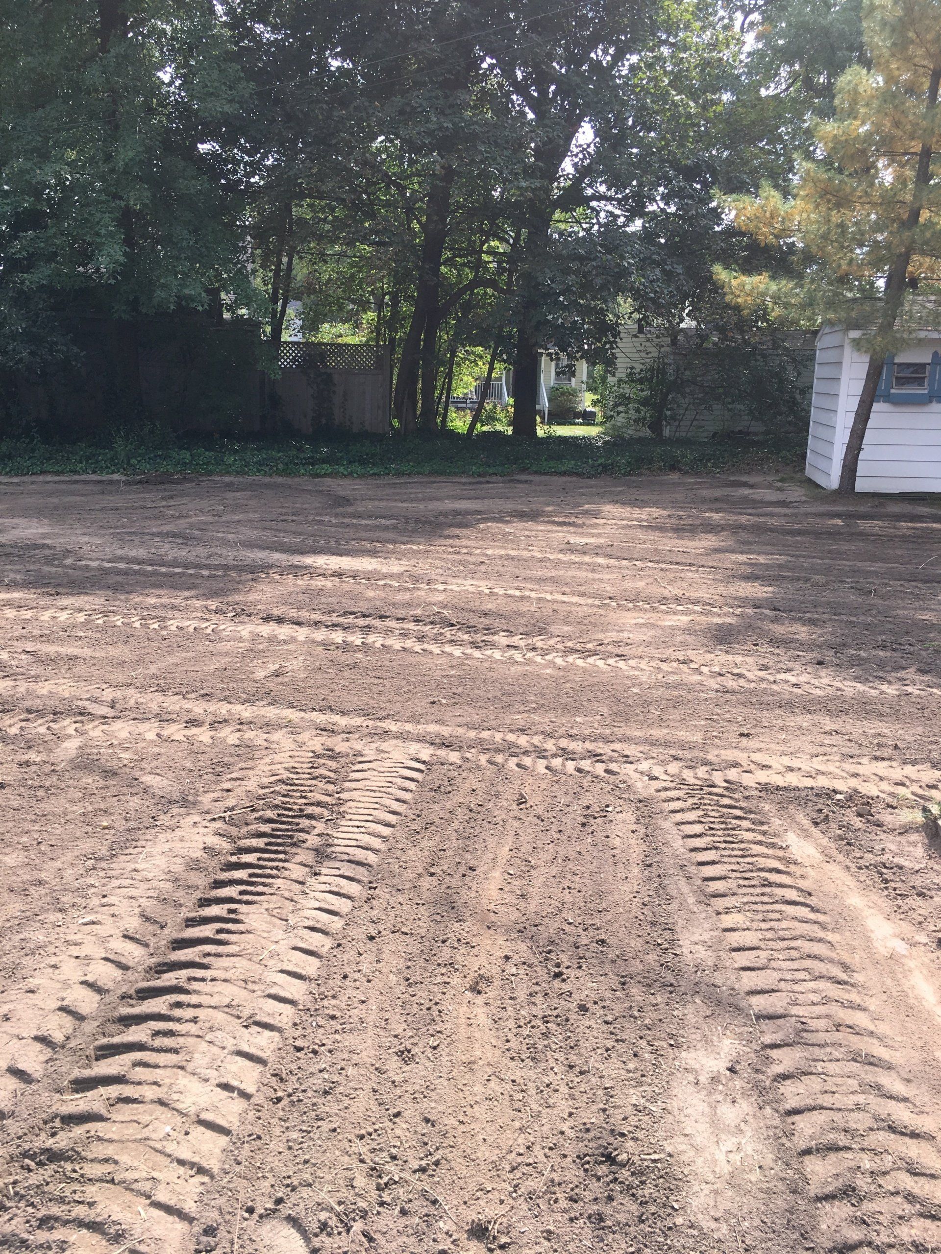 Freshly graded dirt lot with tire tracks, bordered by trees and a small white shed on the right.