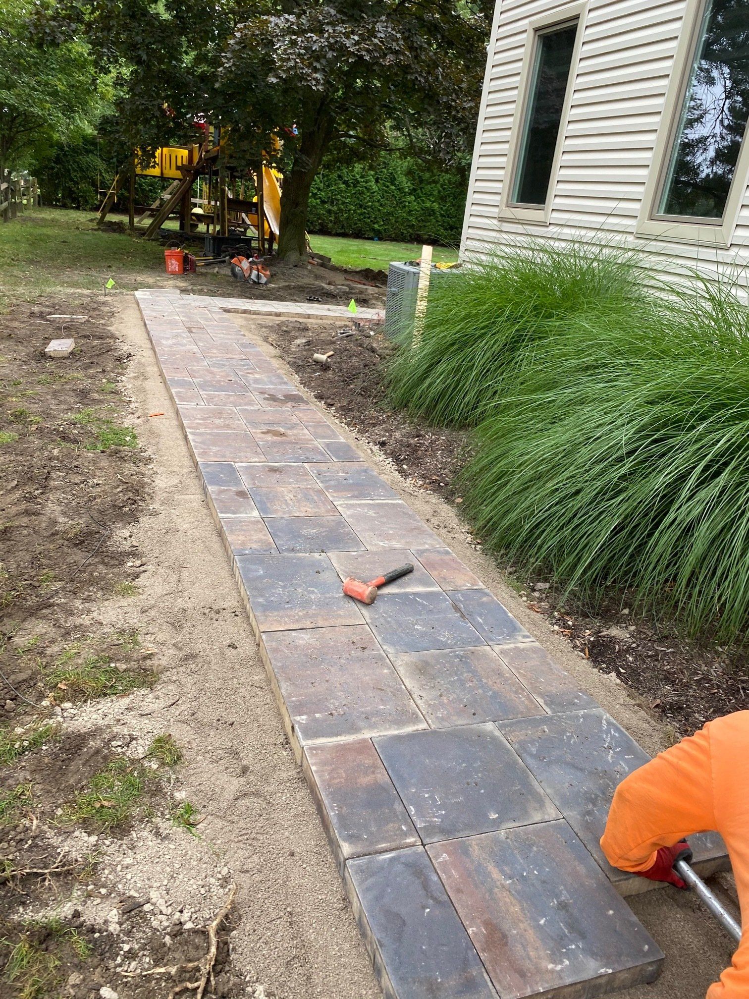 Brick walkway under construction beside a house, with a worker in orange crouching at the edge.